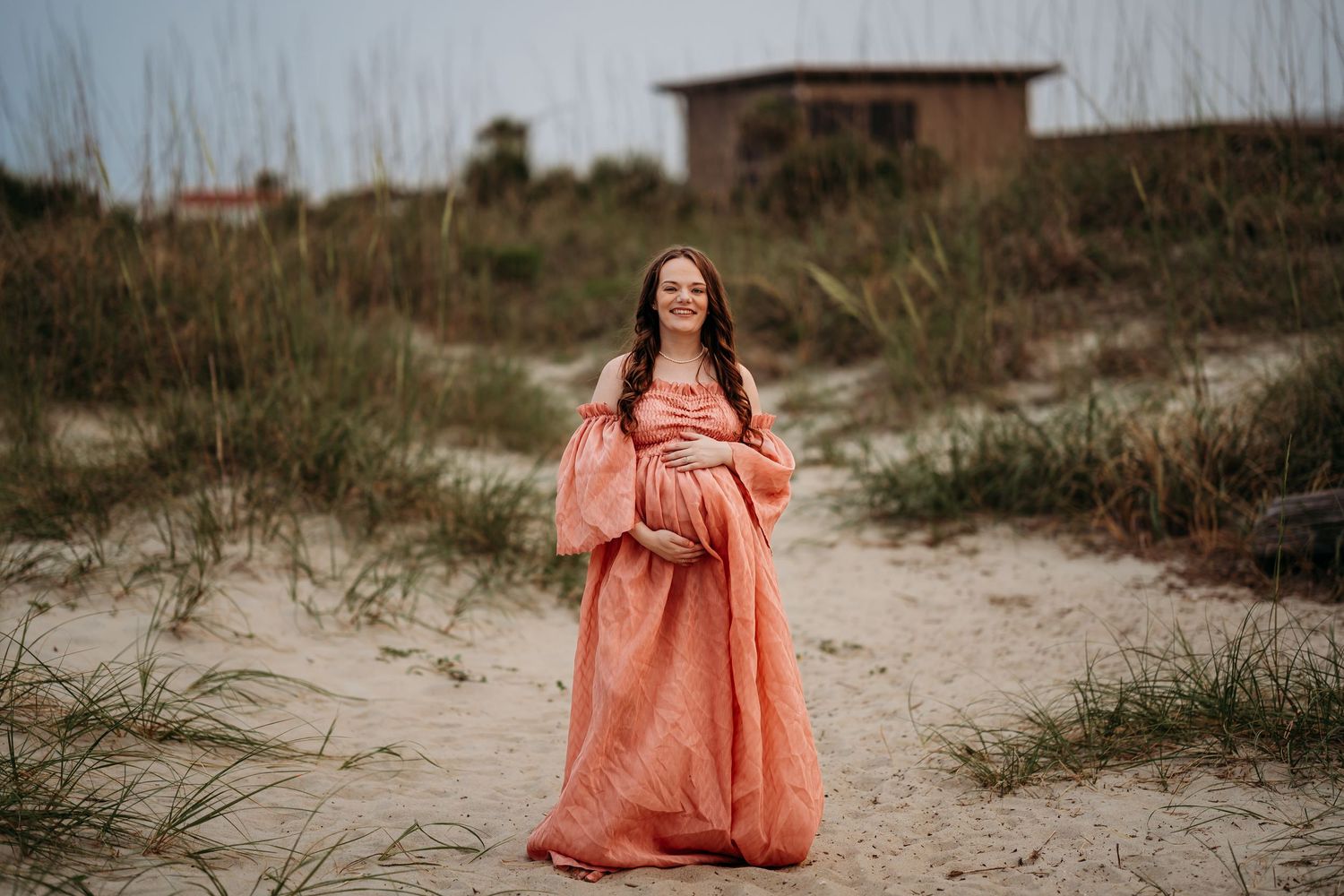 Pregnant woman in flowing coral gown standing on sandy beach path surrounded by dune grass.