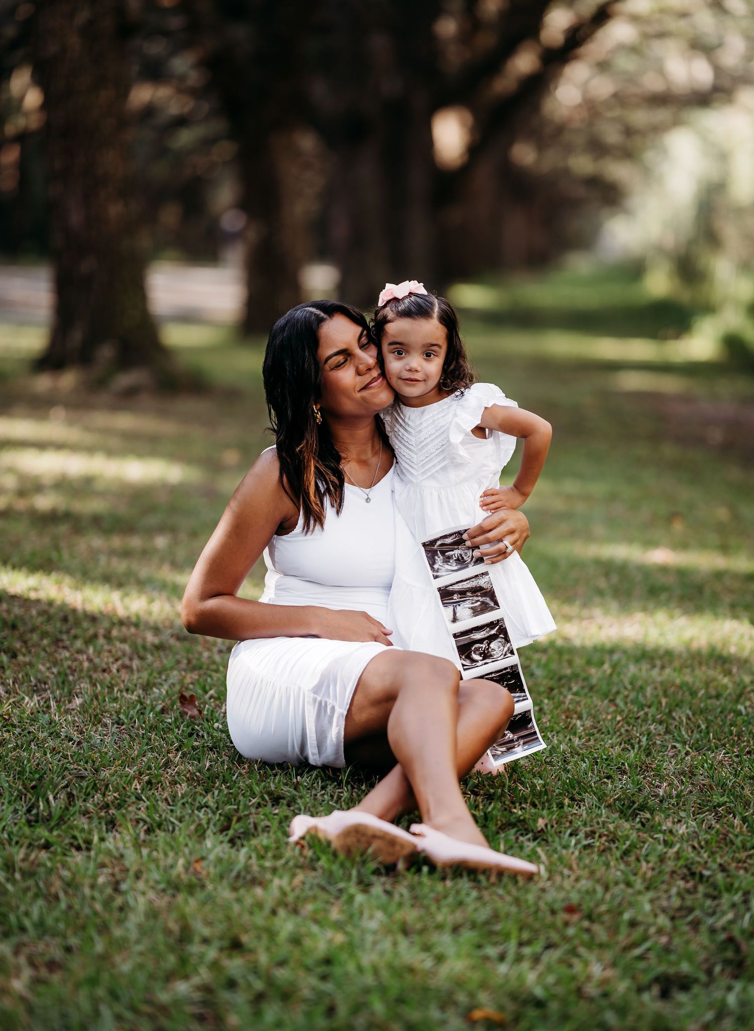 Pregnant woman in white dress sits on grass with toddler daughter holding ultrasound photos in a park.