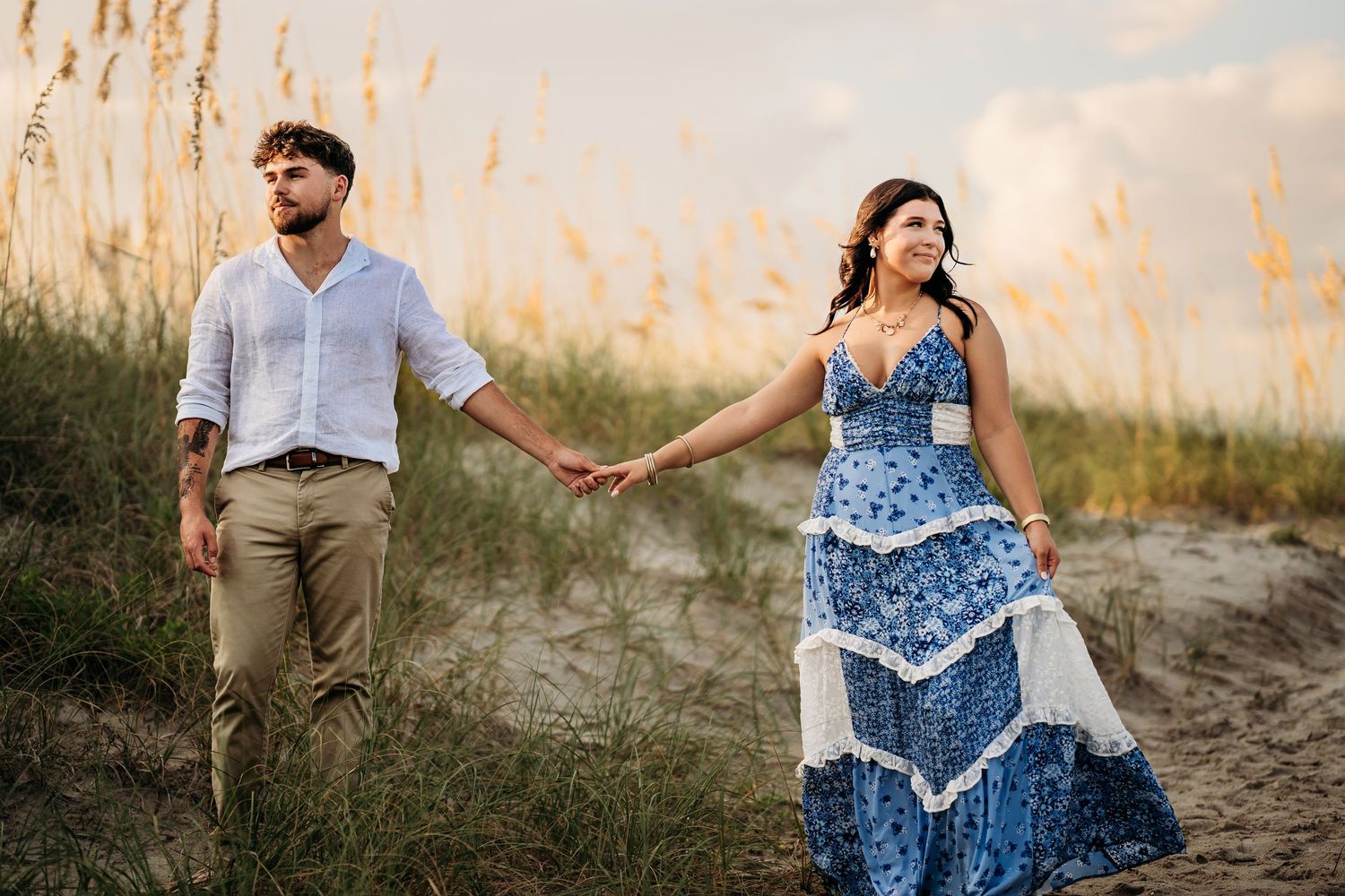 Pregnant couple holding hands walking on beach, woman in blue floral dress, man in white shirt and khaki pants.