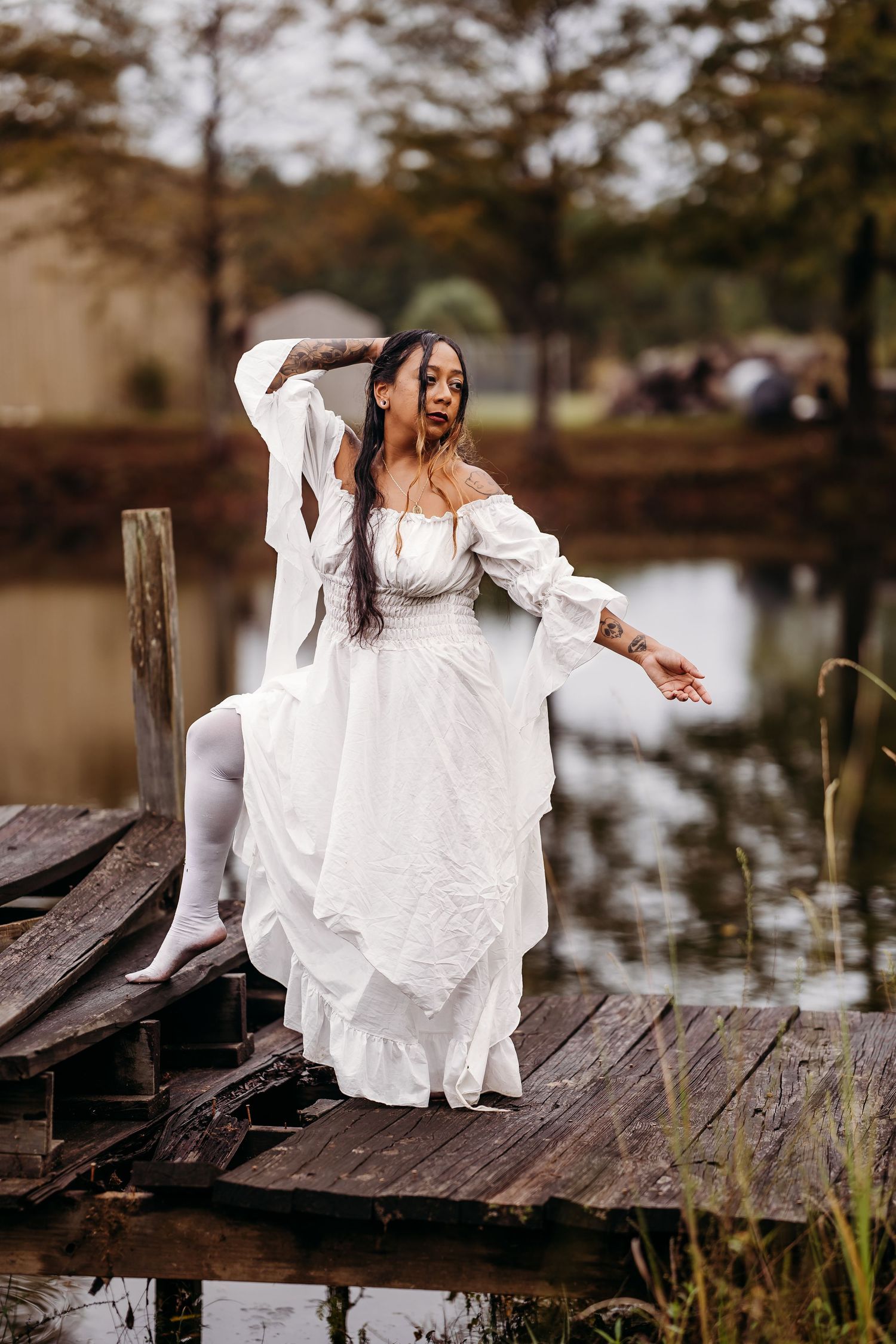 Woman in white off-shoulder dress and white tights poses on rustic wooden dock by calm lake.