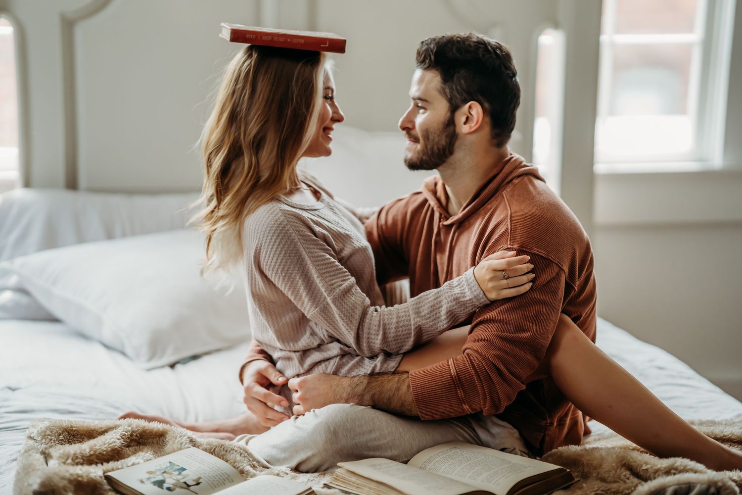 Loving couple sitting face to face on bed with book balanced on head.