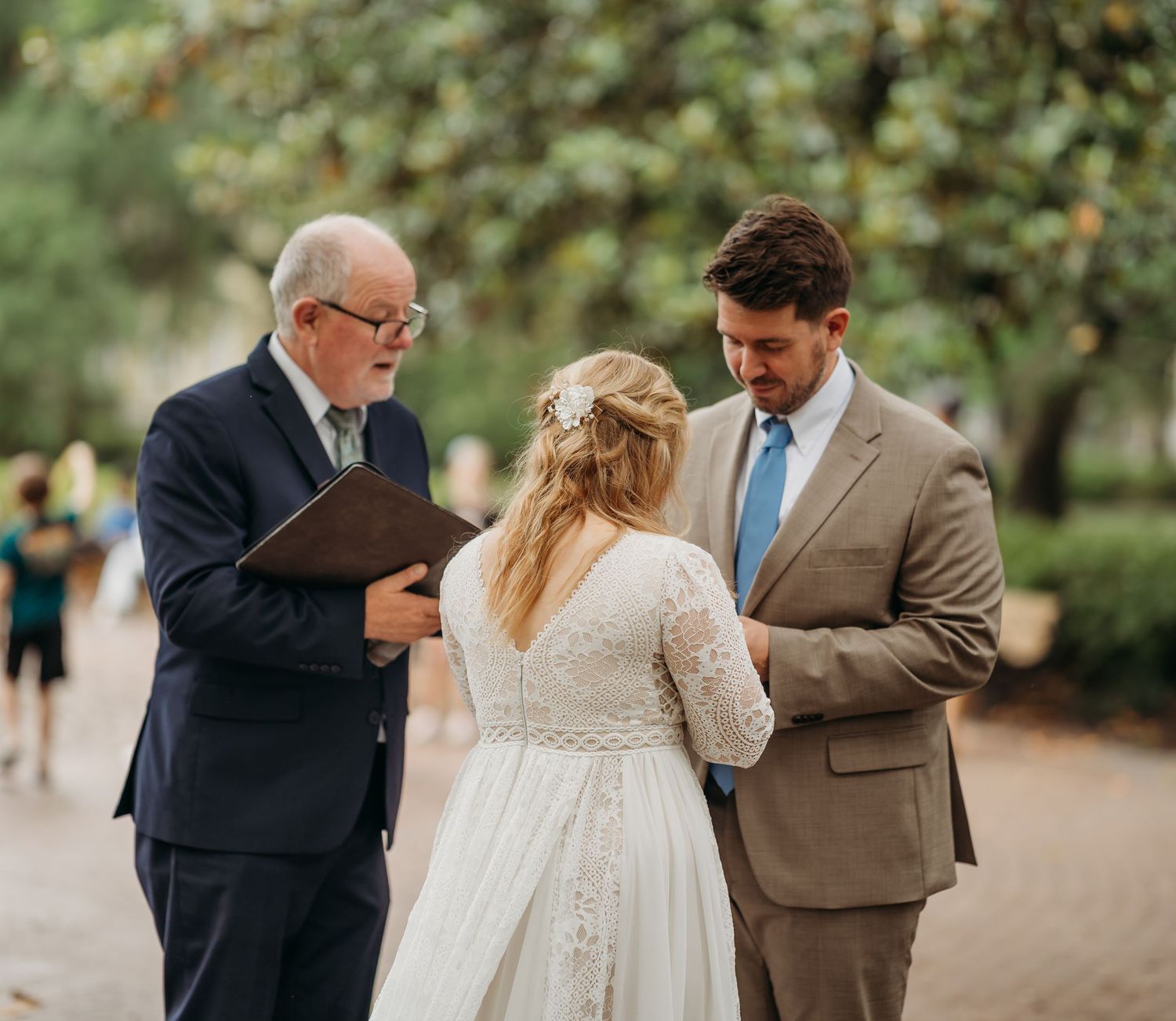 Bride and groom bow heads in prayer with officiant during intimate outdoor wedding ceremony in a lush park setting.