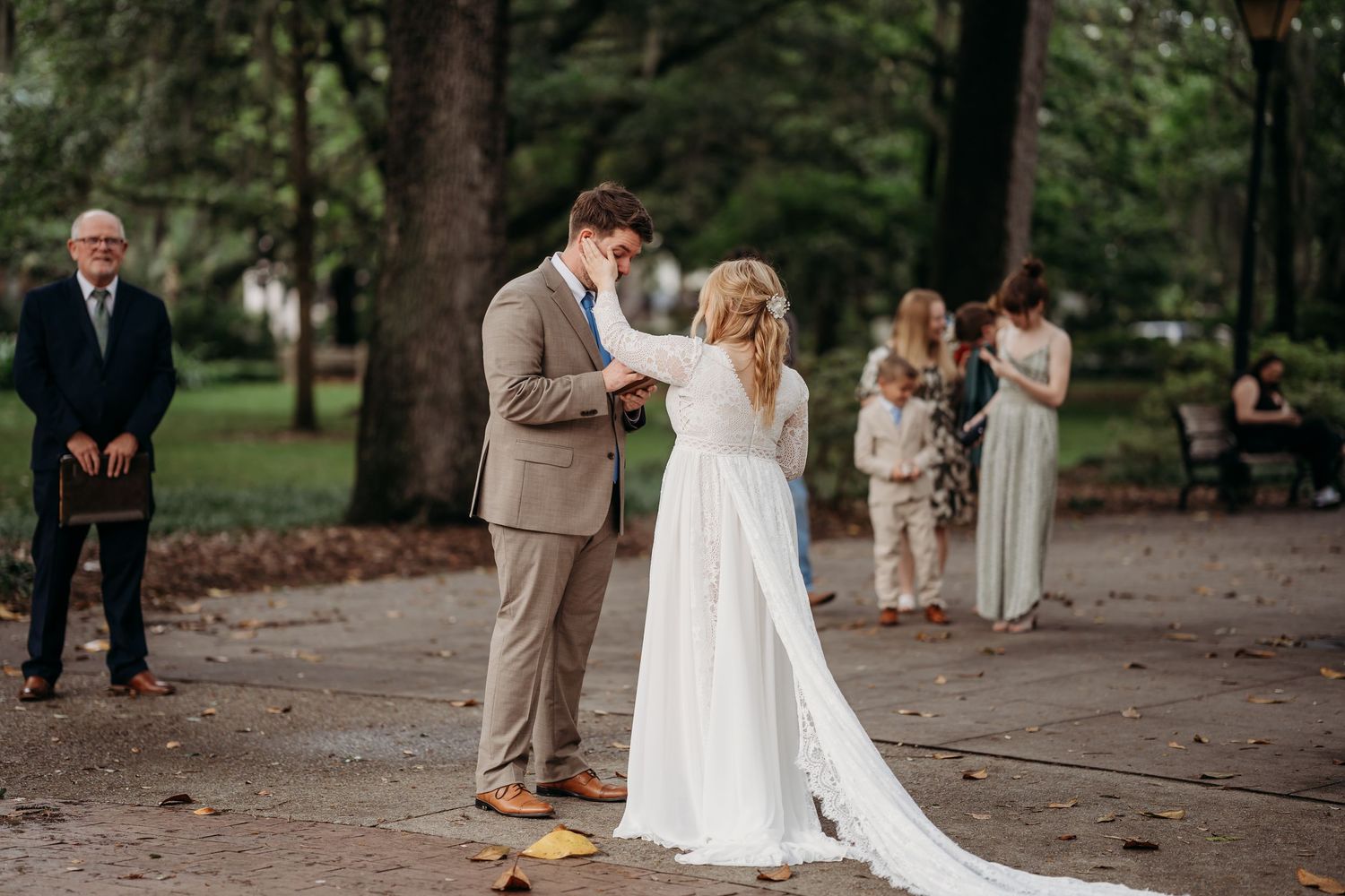 Bride in white lace gown and groom in tan suit exchange vows at outdoor park wedding ceremony.