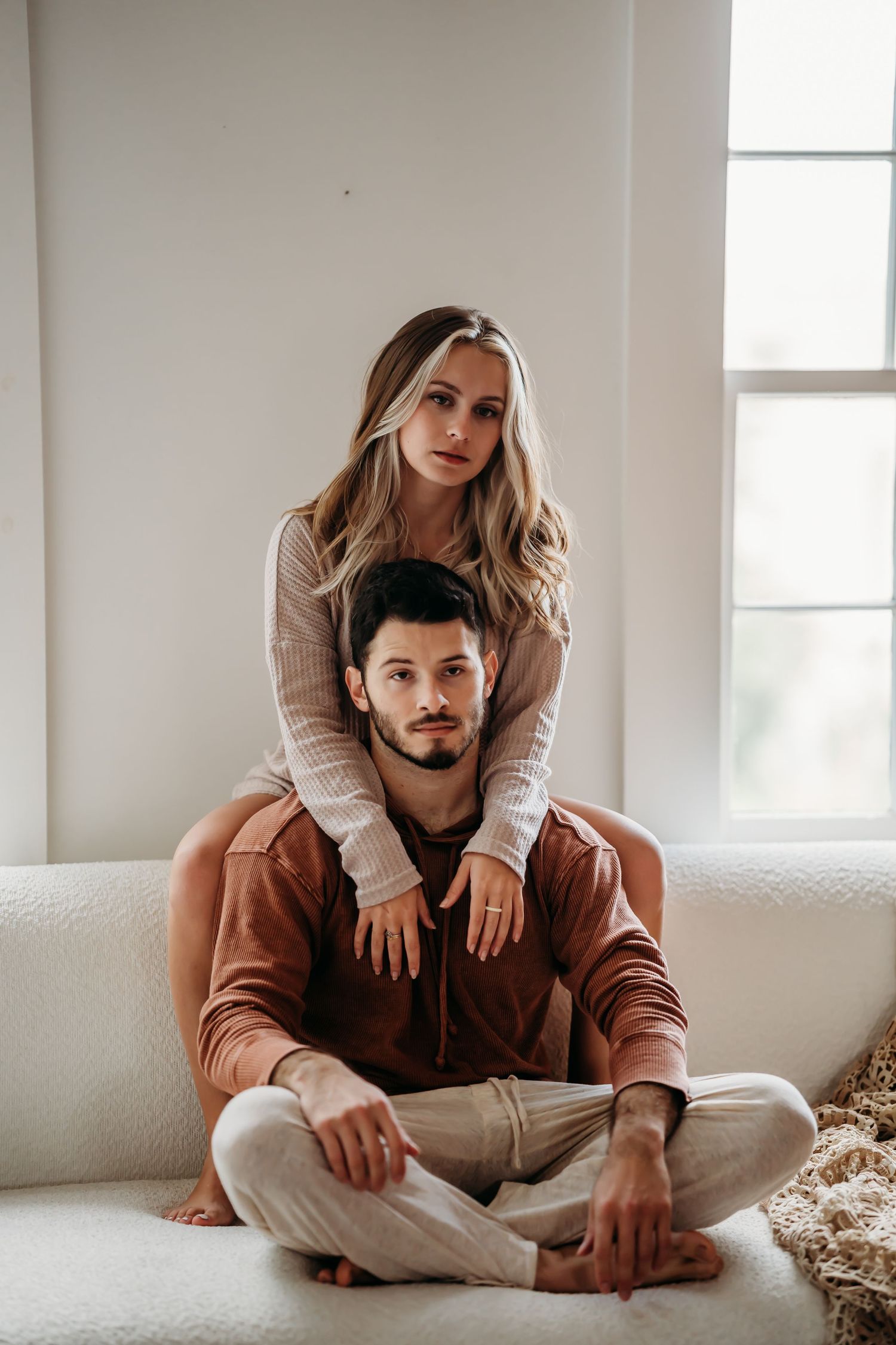 A stylish couple posing together, the woman sitting behind the man on a white sofa near a window.