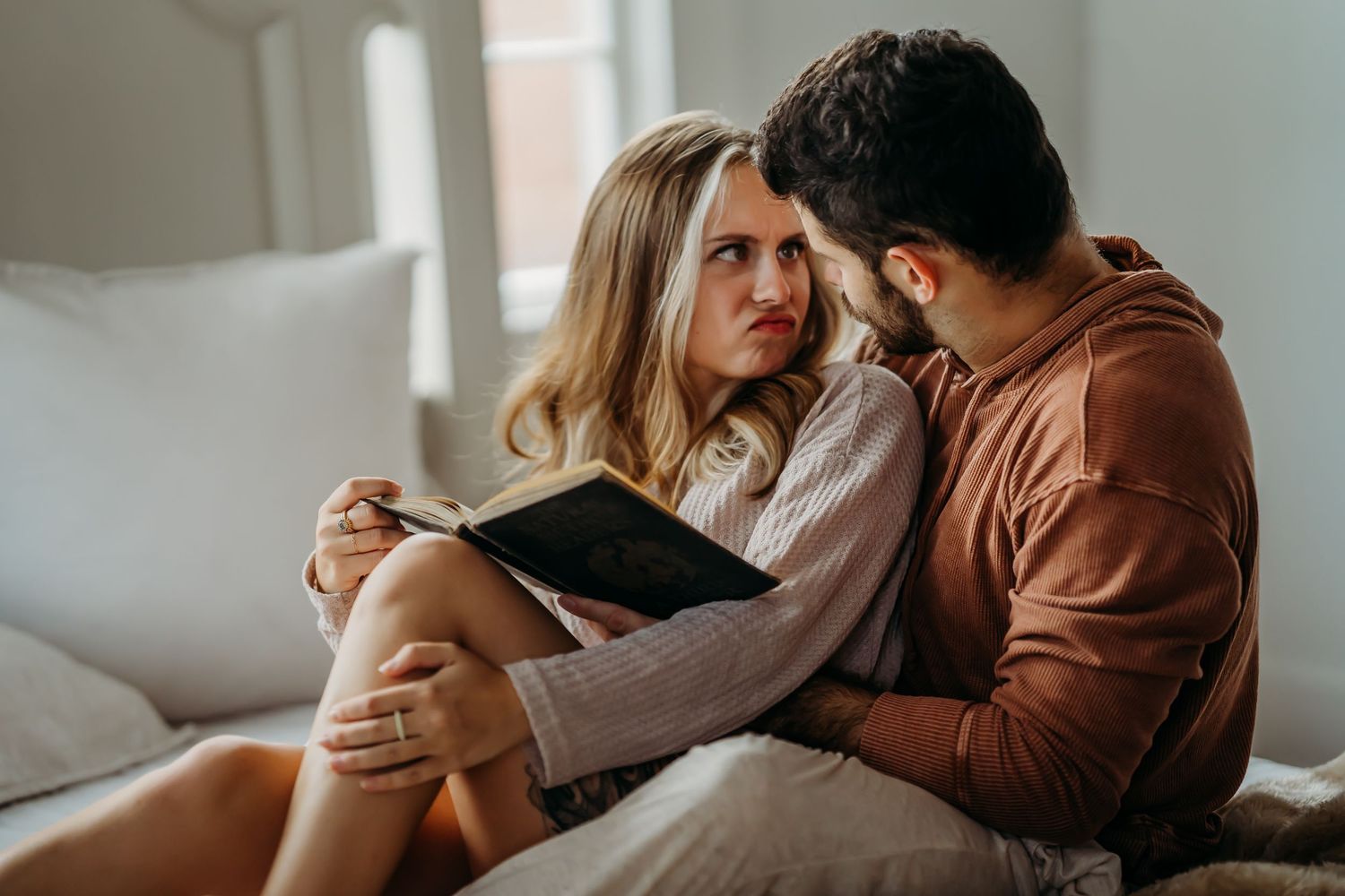 A couple sits on a bed as the woman holds a book and gazes at the man beside her.
