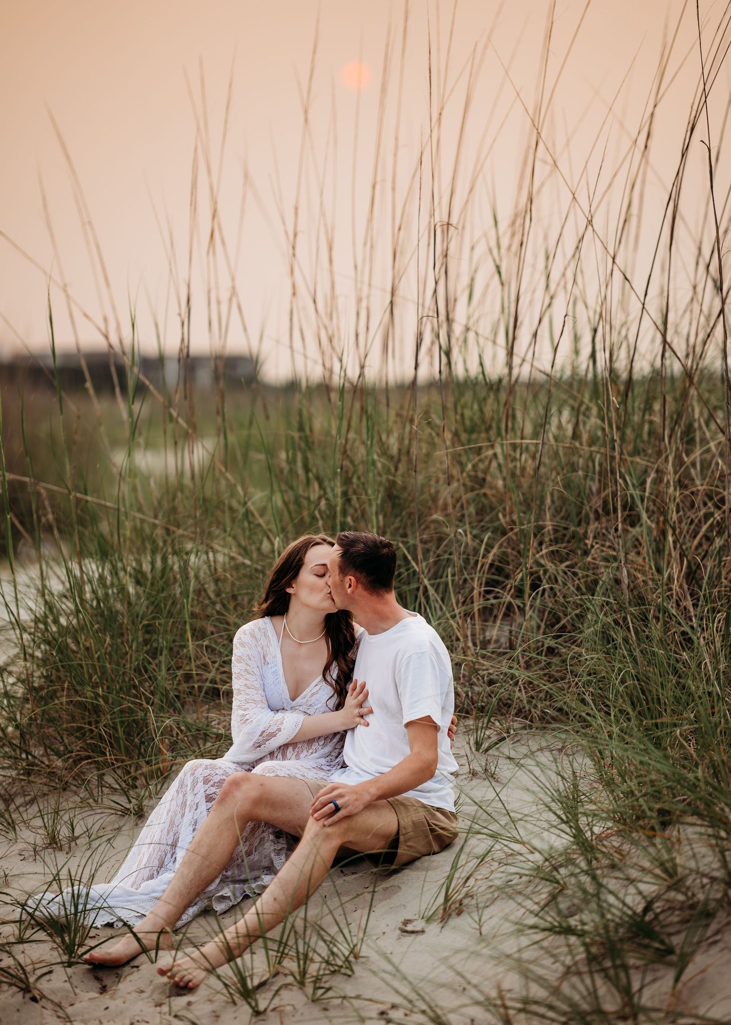 Couple sharing a kiss while sitting in beach sand dunes surrounded by tall sea grass at sunset.