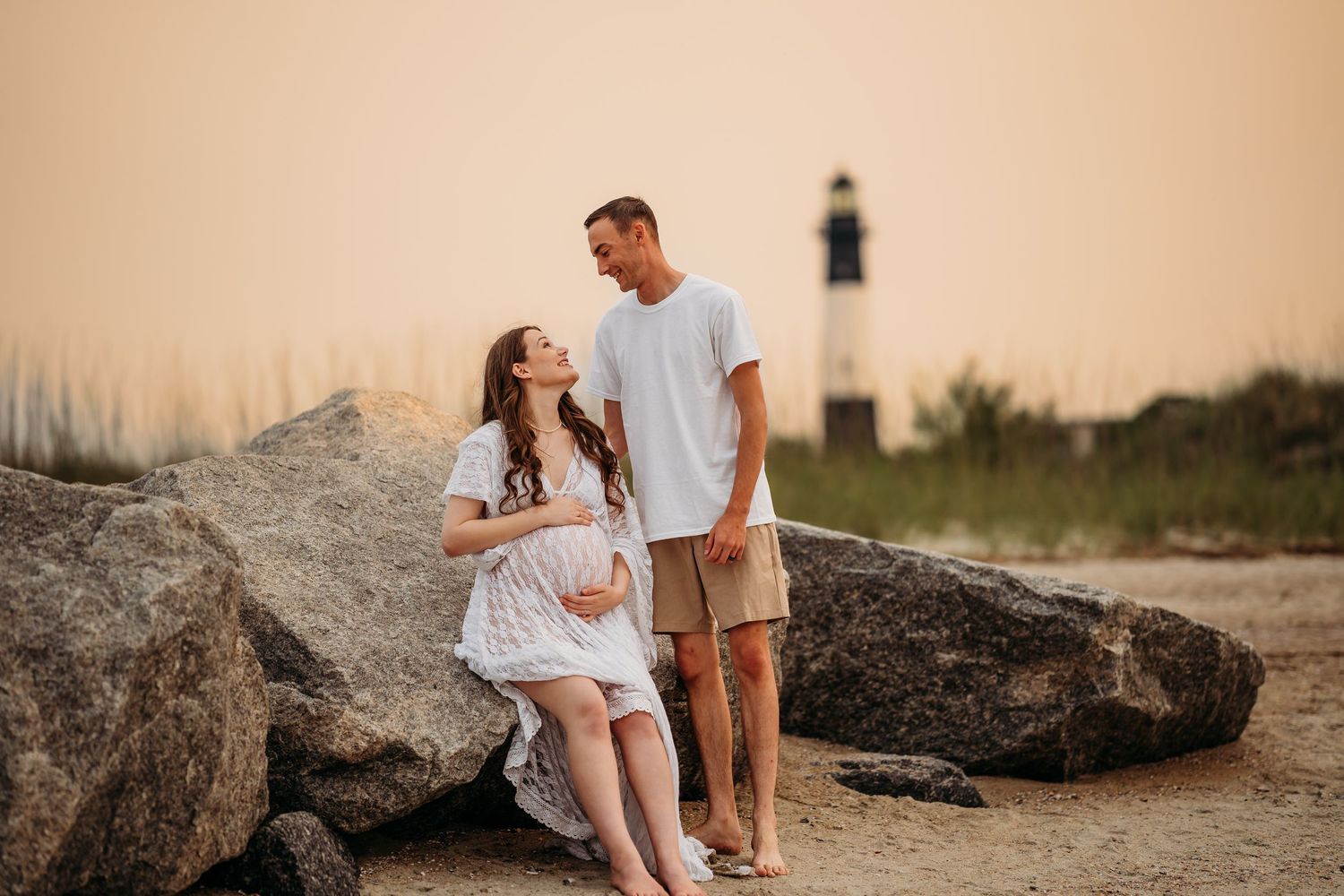Pregnant couple sharing a tender moment near large rocks on beach with lighthouse in golden sunset background.