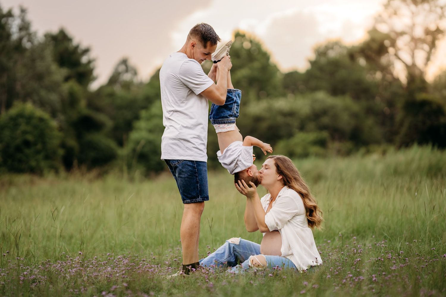 Family playing outdoors in a green meadow at sunset, father lifting child while mother sits smiling on the grass.