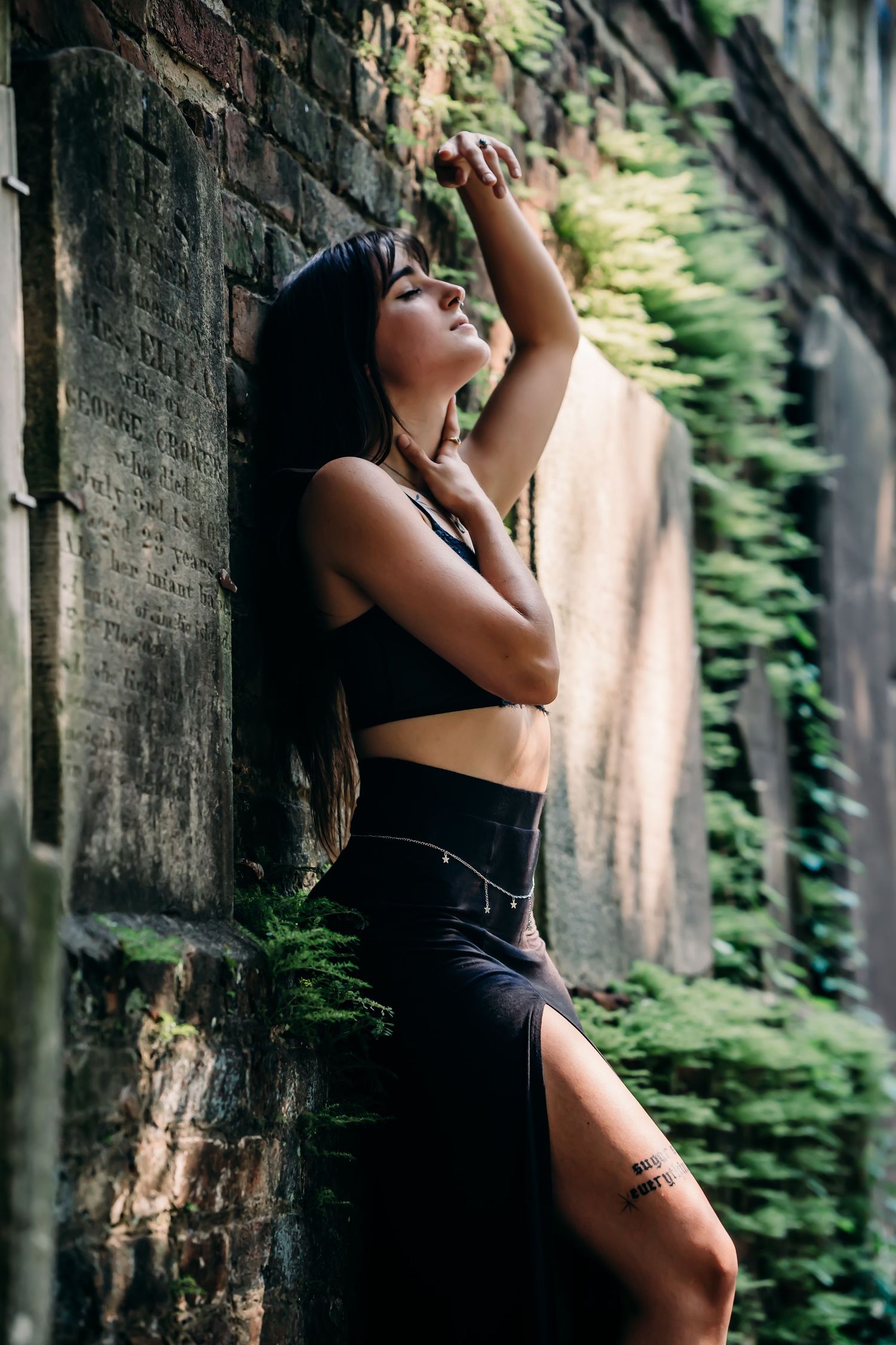 Woman in black crop top and slit skirt poses against mossy stone wall surrounded by lush green ferns.