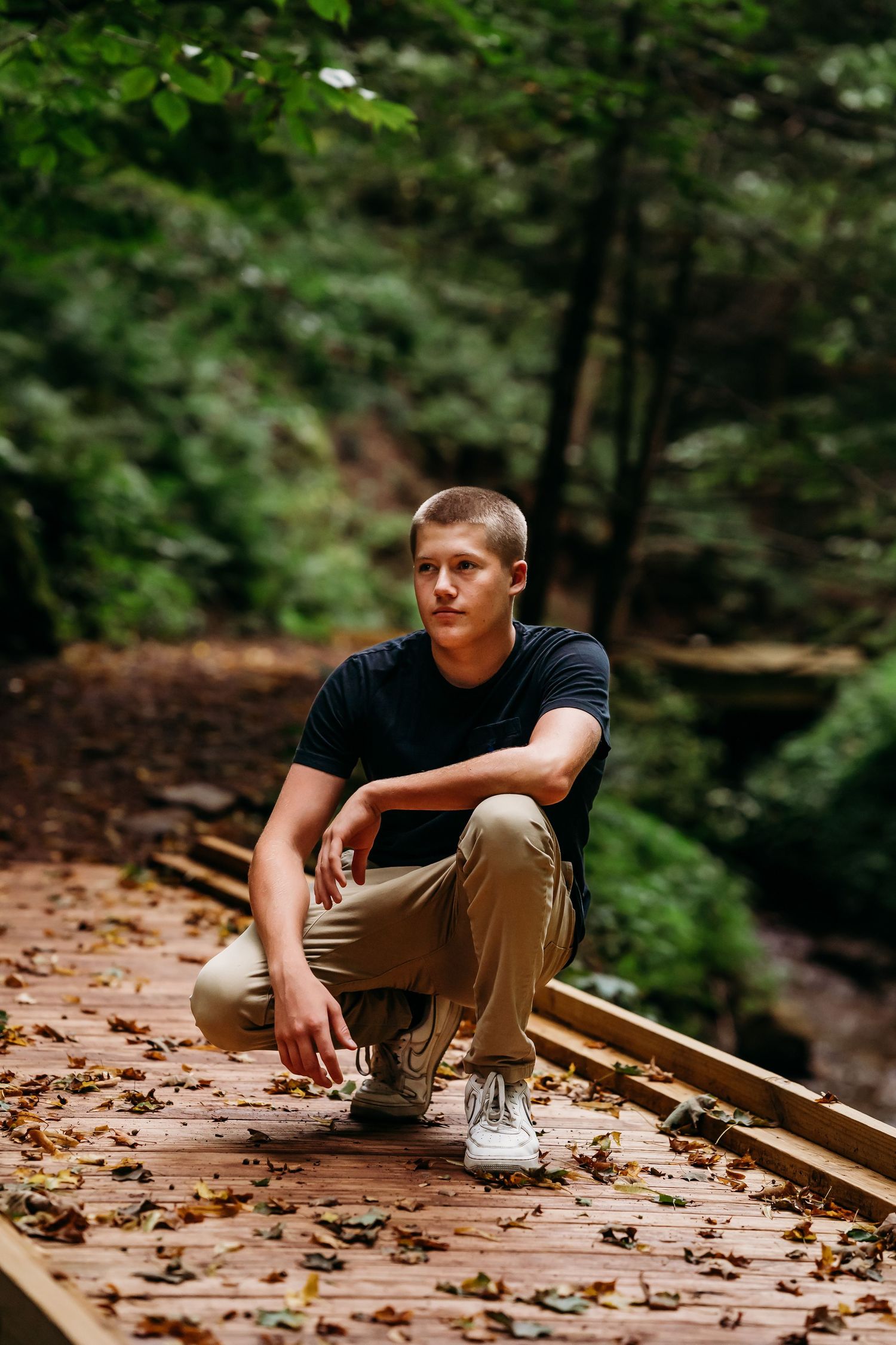 Young man in black t-shirt and khaki pants crouching on a wooden boardwalk trail surrounded by lush green forest.