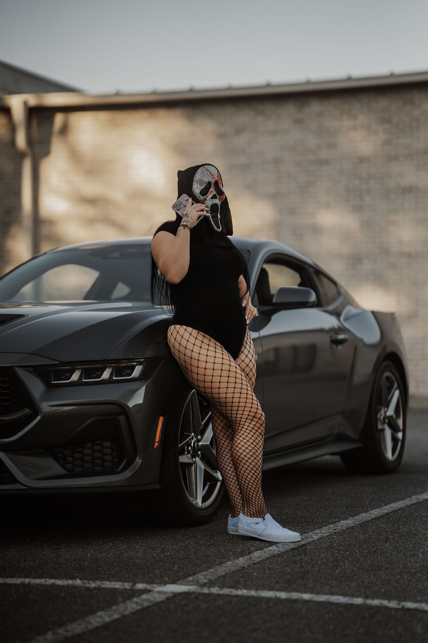 Person in Scream mask and fishnet tights leaning against a gray Ford Mustang in a parking lot.