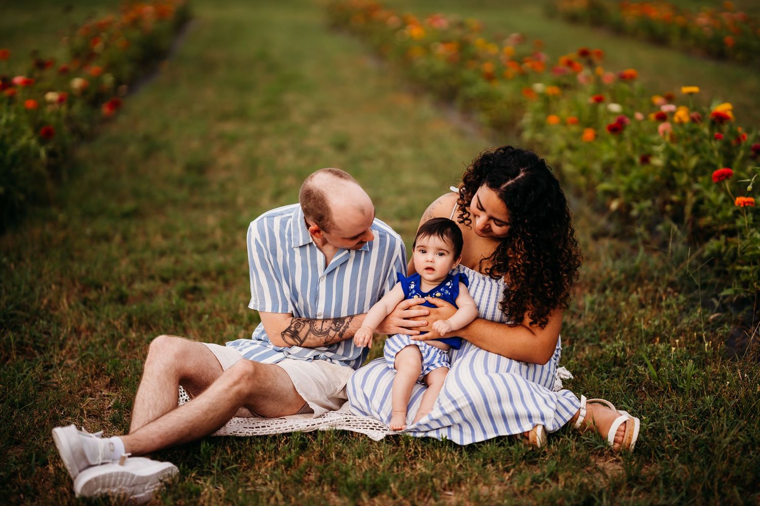 Family of three sitting on a blanket in a flower field, parents lovingly gazing at their baby boy.