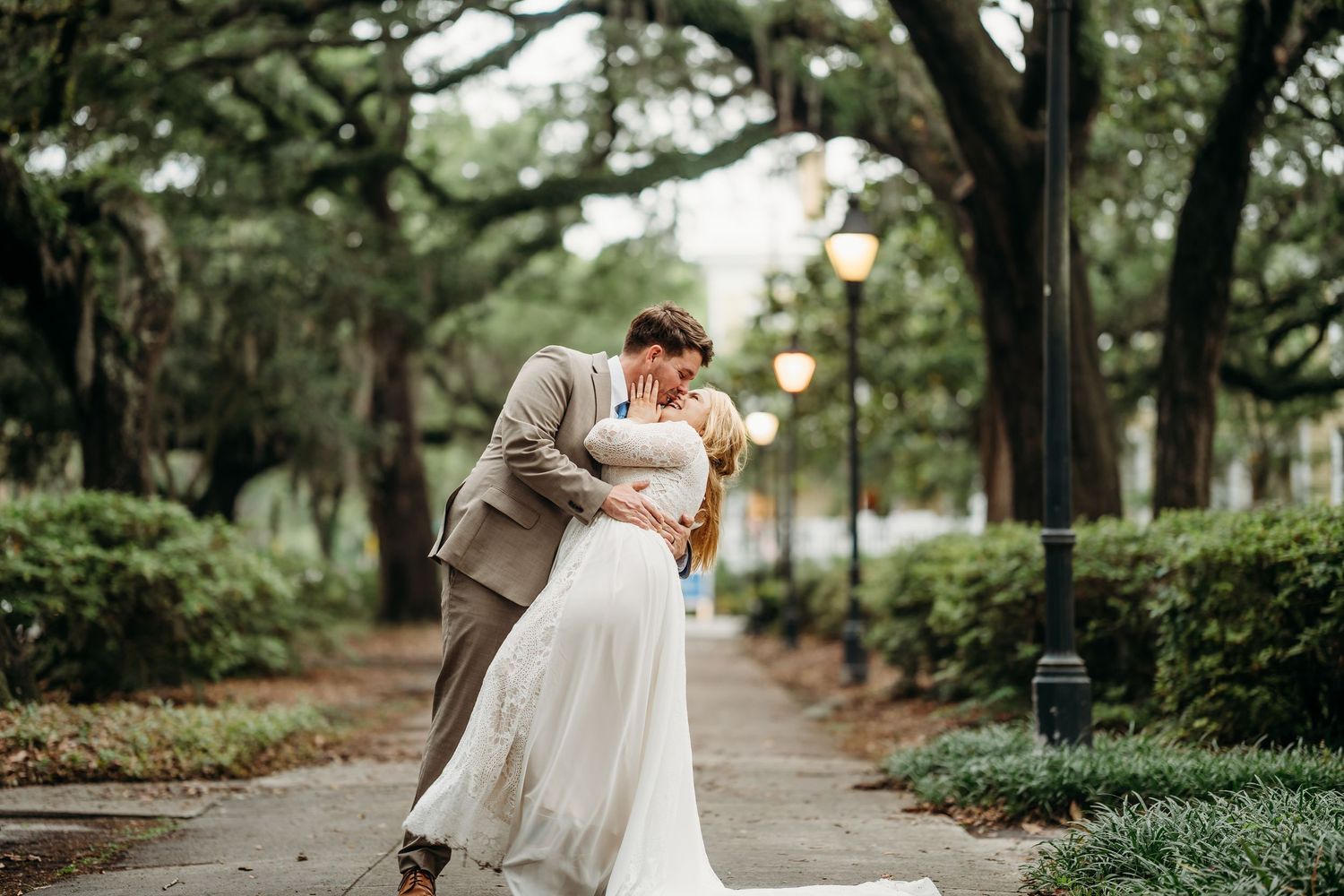 Groom dips his bride for a passionate kiss along a beautiful moss-draped tree-lined walkway.