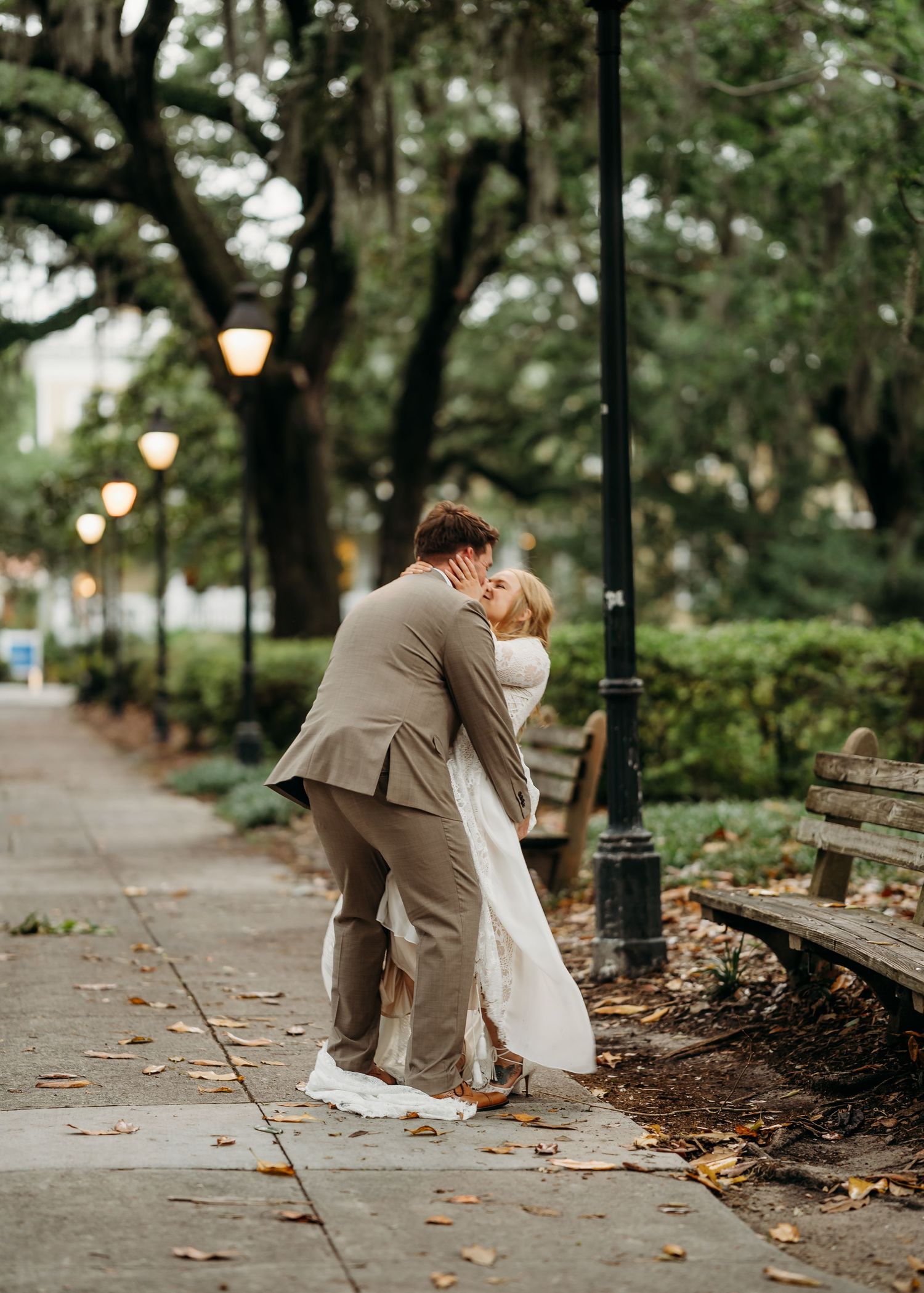 Bride and groom share a sweet kiss beside a vintage lamppost on a leaf-strewn garden path.