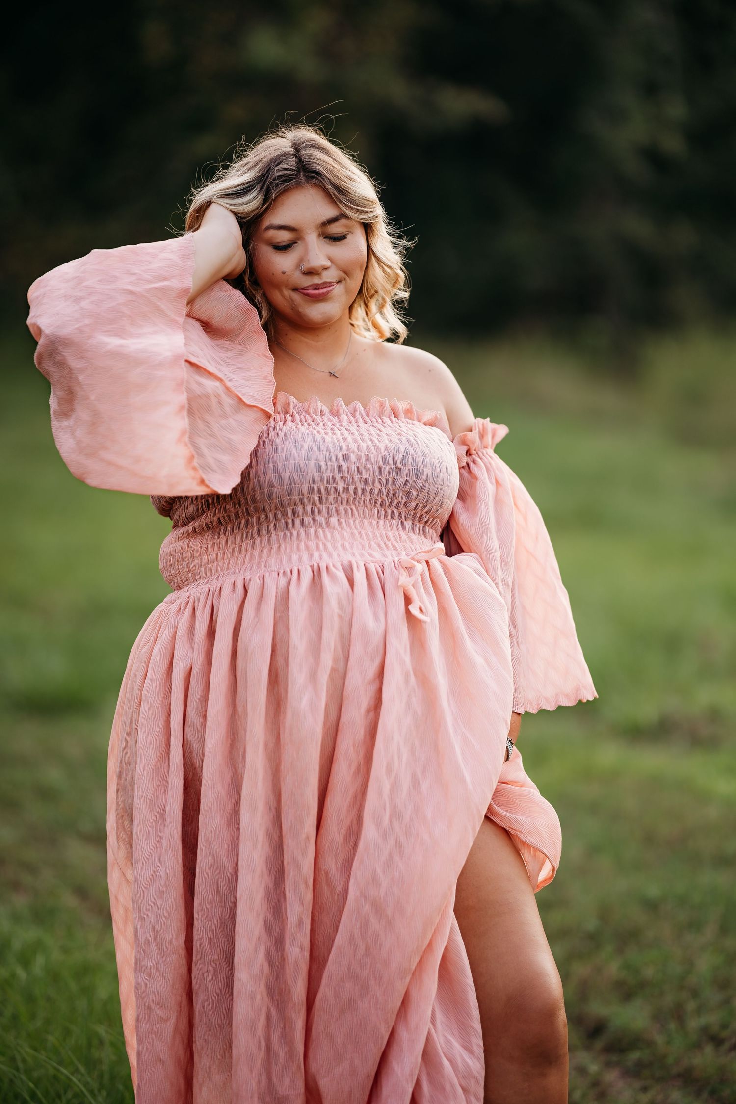 Beautiful blonde woman in an elegant off-shoulder pink maxi dress posing outdoors in a field.