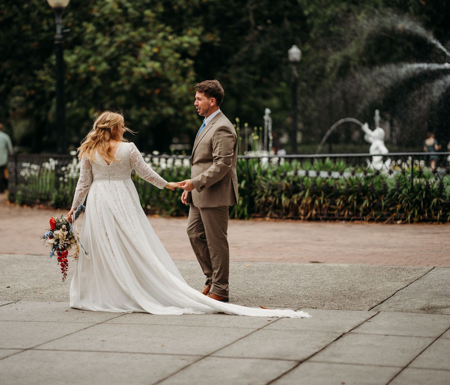 Bride in flowing white gown and groom walk hand-in-hand near a park fountain.