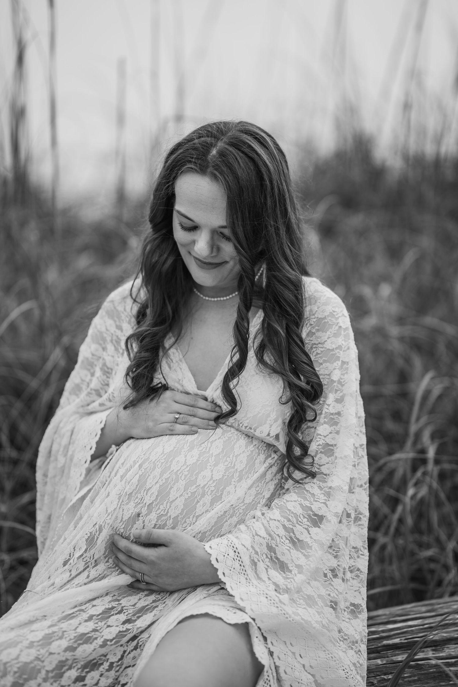 Black and white portrait of pregnant woman in lace gown gently cradling her baby bump outdoors.
