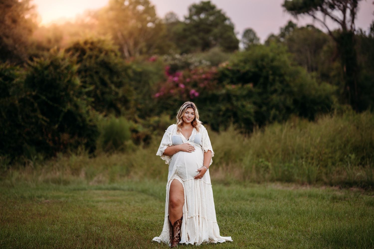 Pregnant woman in a flowing white maternity gown standing in a sunlit green field at golden hour.