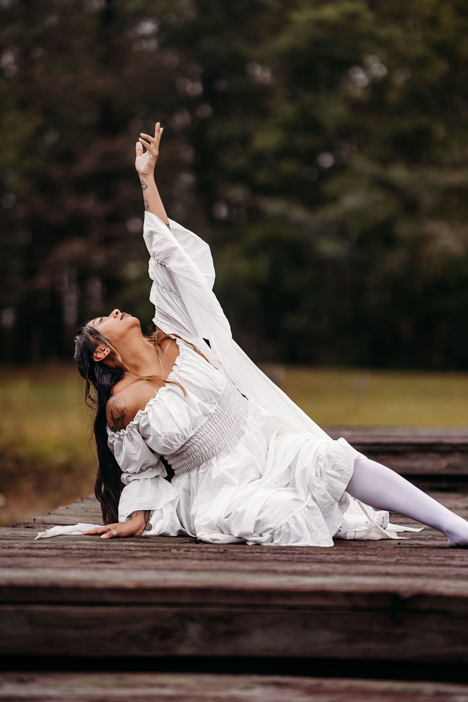 Woman in white dress performing expressive dance pose on wooden deck with arm raised.