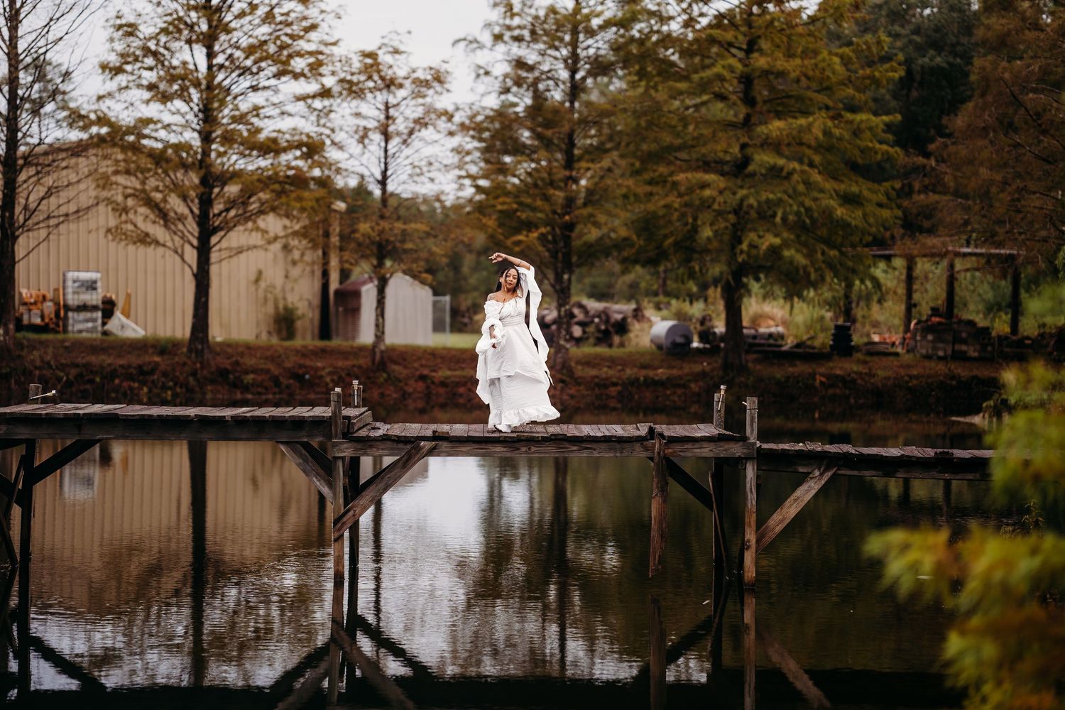 Bride in elegant white wedding dress standing on rustic wooden dock beside tranquil pond with fall trees.
