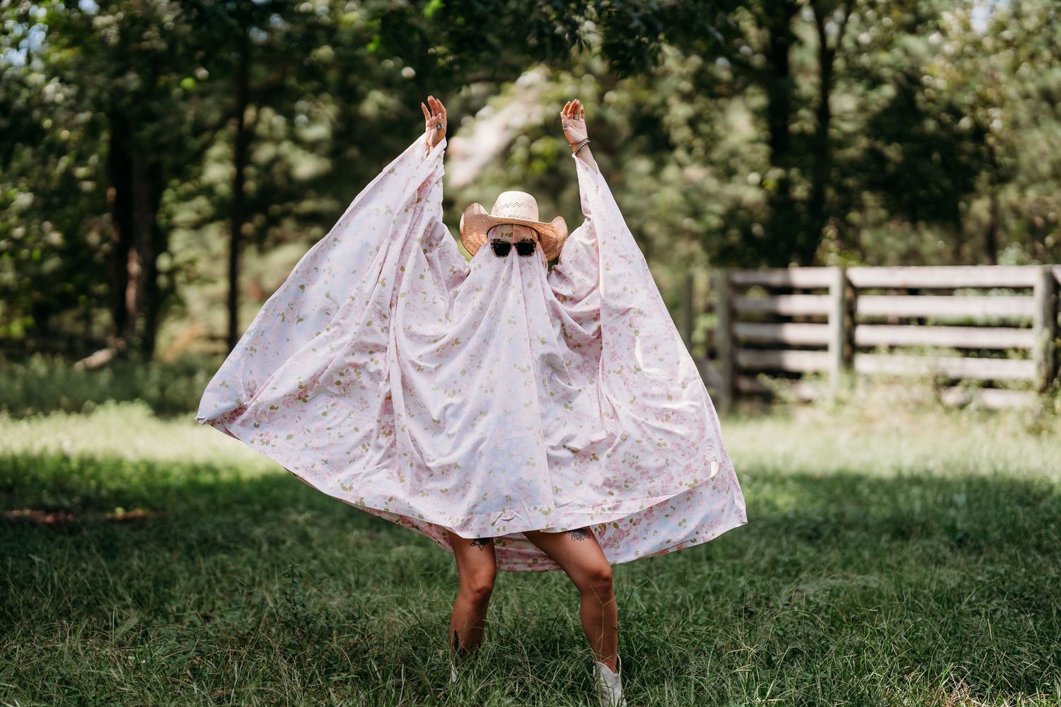 Woman in white lace dress and hat spreading arms wide open in green field.