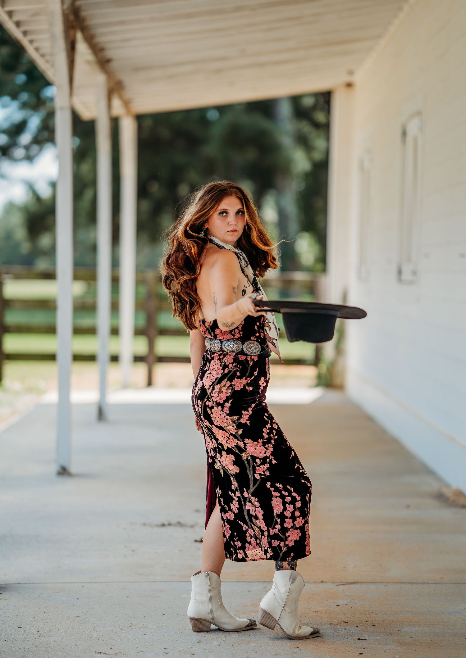 Woman in floral midi dress and white cowboy boots holding black hat on rustic farm porch.