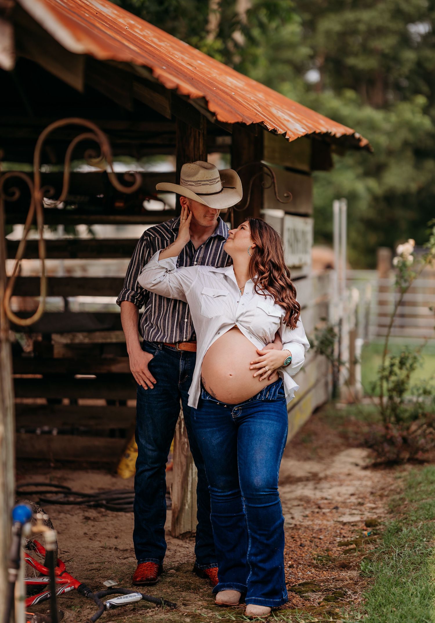 Couple in country attire poses outdoors, woman showing pregnant belly, man in cowboy hat embracing her near rustic barn.