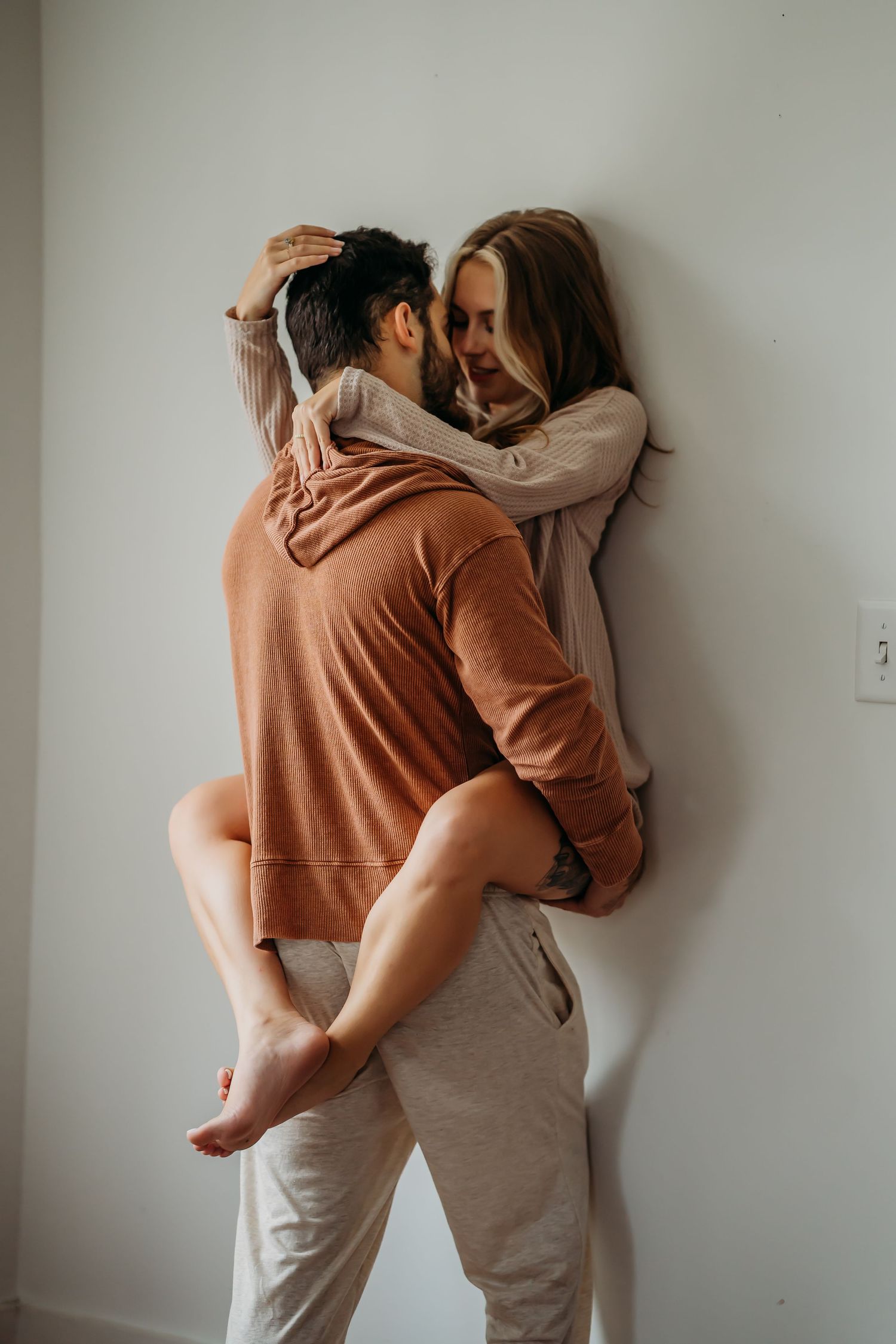 A man lifts a smiling woman against a white wall as they share a joyful embrace.
