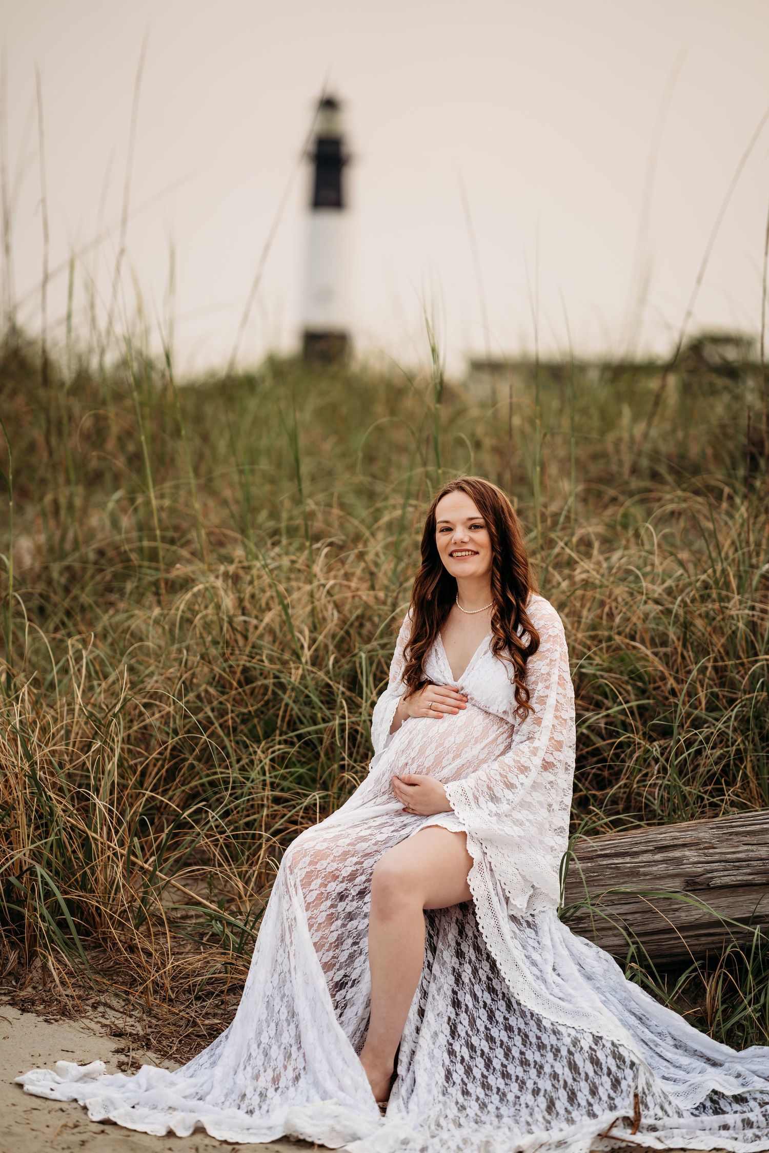 Pregnant woman in flowing white lace dress sits on driftwood log near coastal lighthouse at sunset.