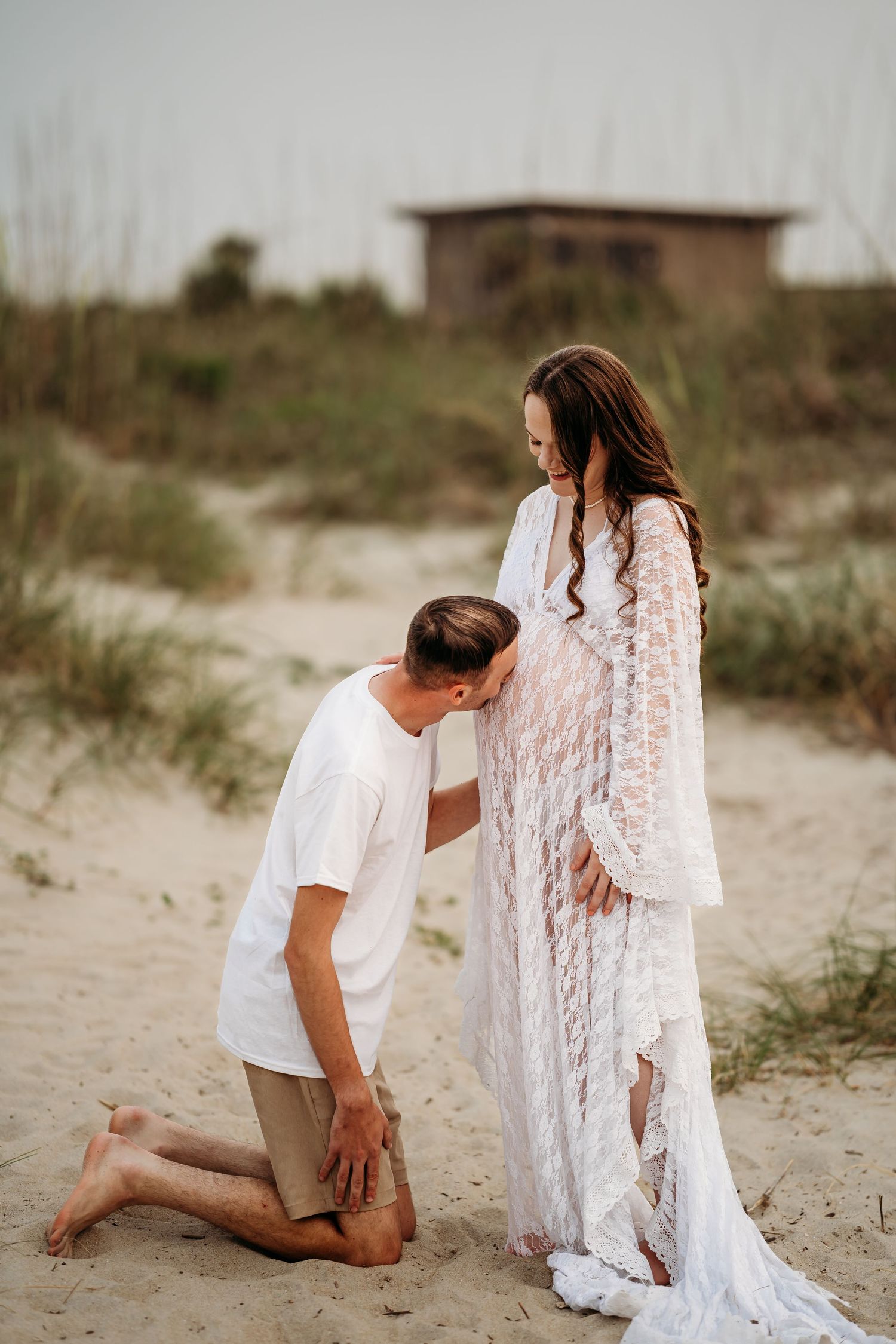 Man kneeling to kiss pregnant woman's baby bump on sandy beach during maternity photo session.
