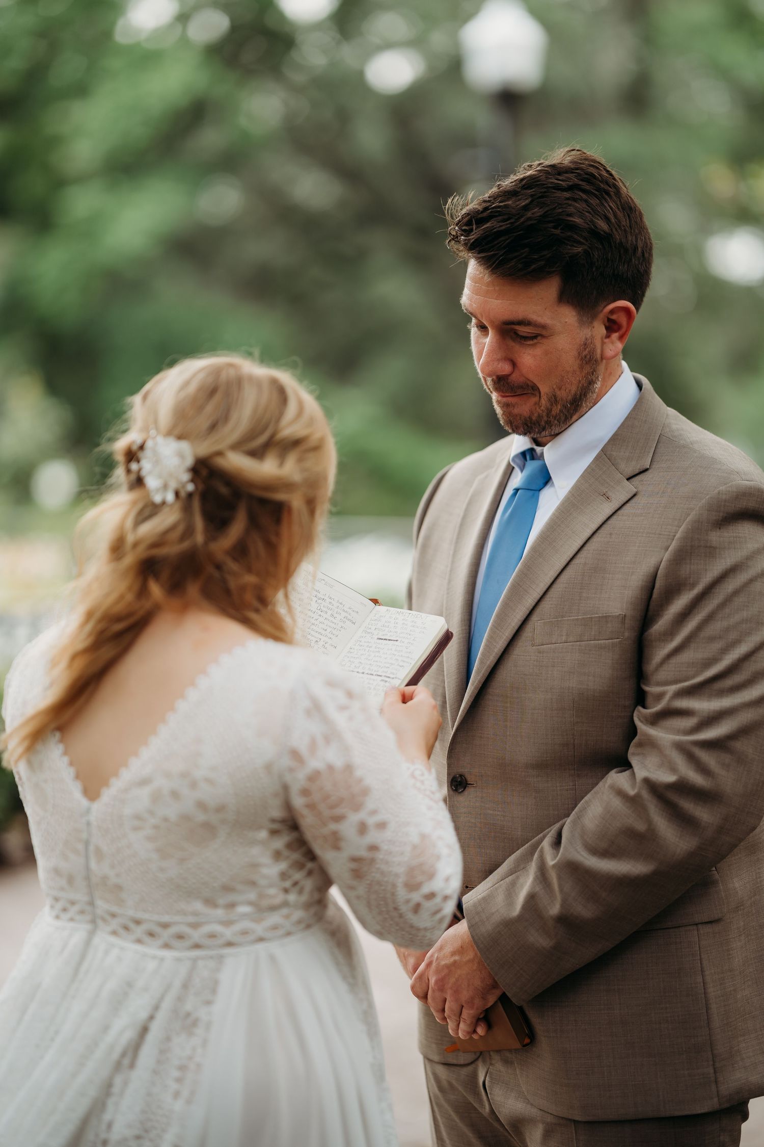 Groom in tan suit listens emotionally as bride reads handwritten vows during outdoor ceremony.