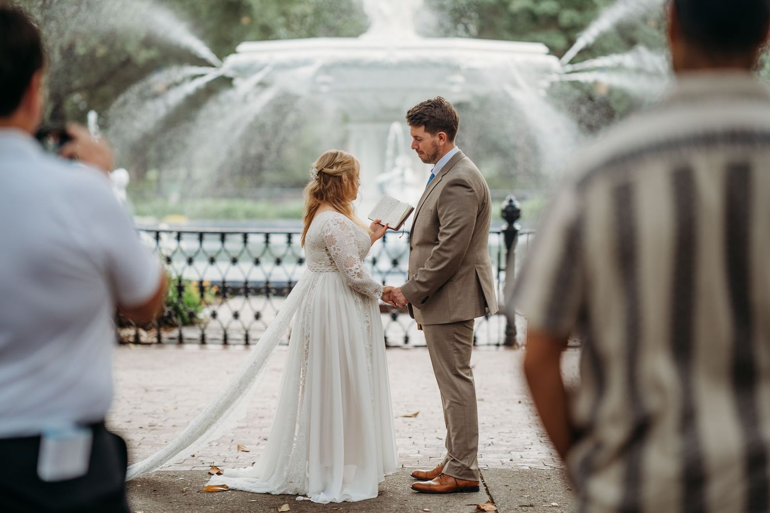 Bride and groom hold hands exchanging vows at outdoor wedding ceremony in front of fountain.