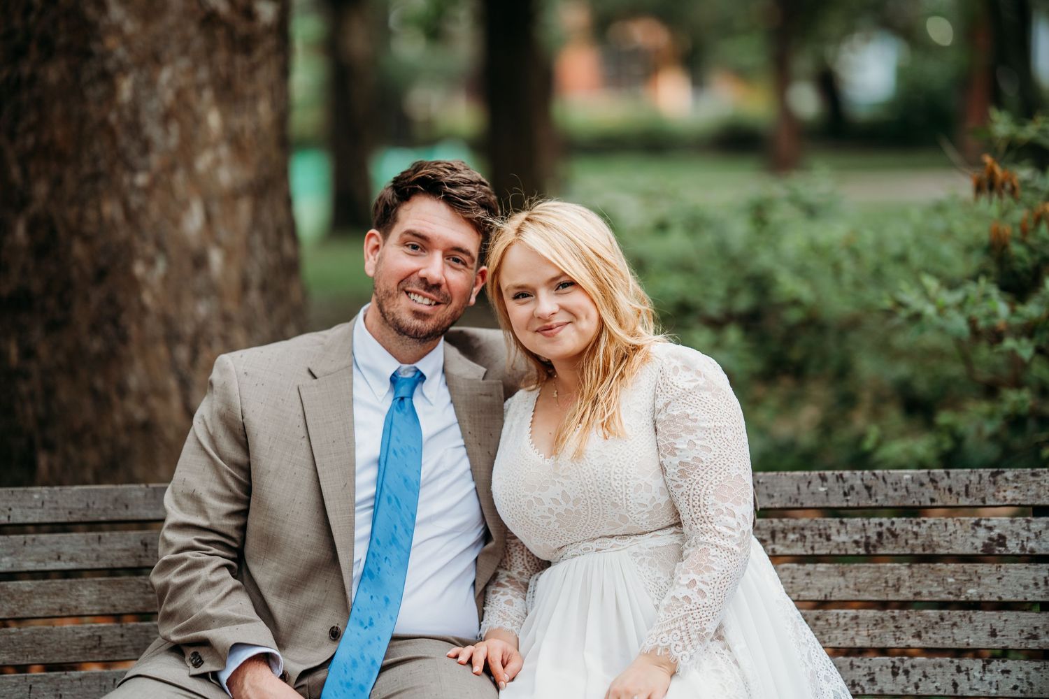 Smiling bride and groom posing together on a park bench, groom wearing a blue tie and tan suit.