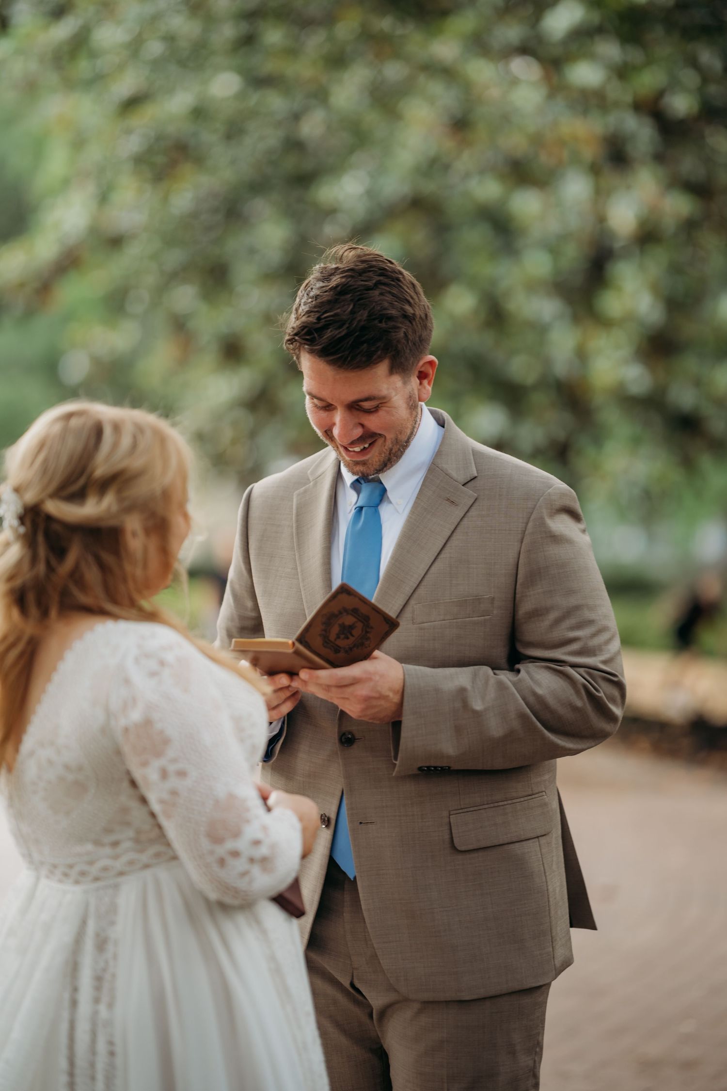 Emotional groom in tan suit reads vows from wooden book holder during outdoor wedding ceremony.