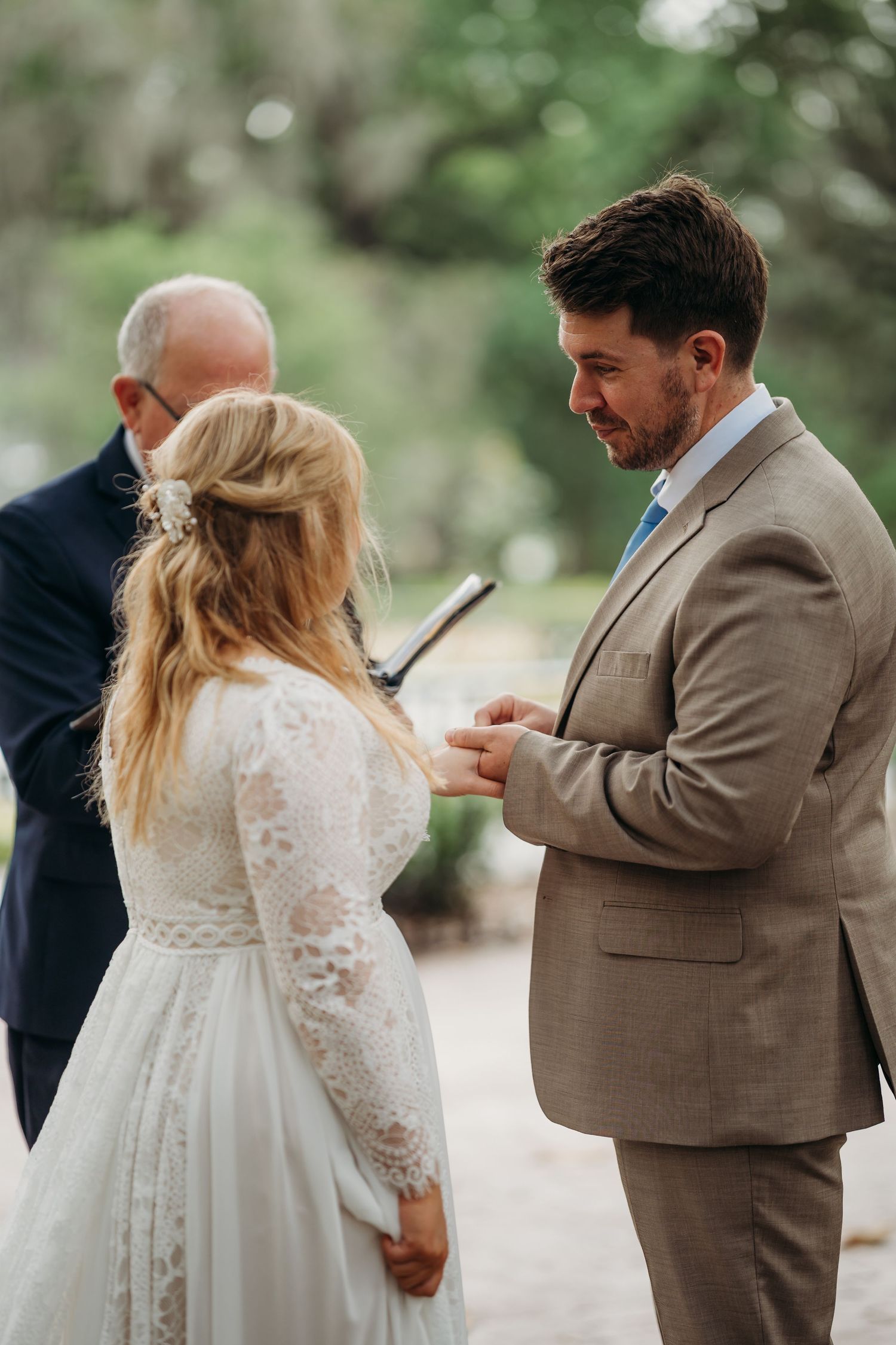 Groom tenderly places wedding ring on bride's finger during an intimate outdoor ceremony.