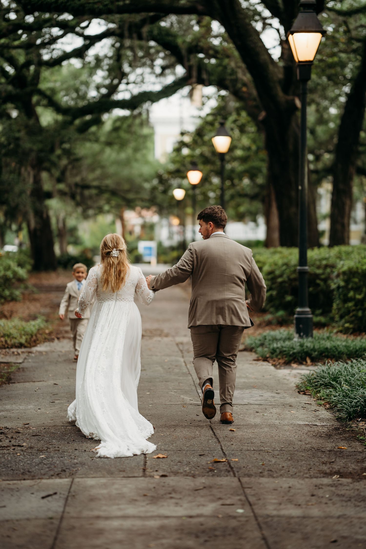 Bride and groom walk hand-in-hand with child ahead on glowing lamp-lit park path.