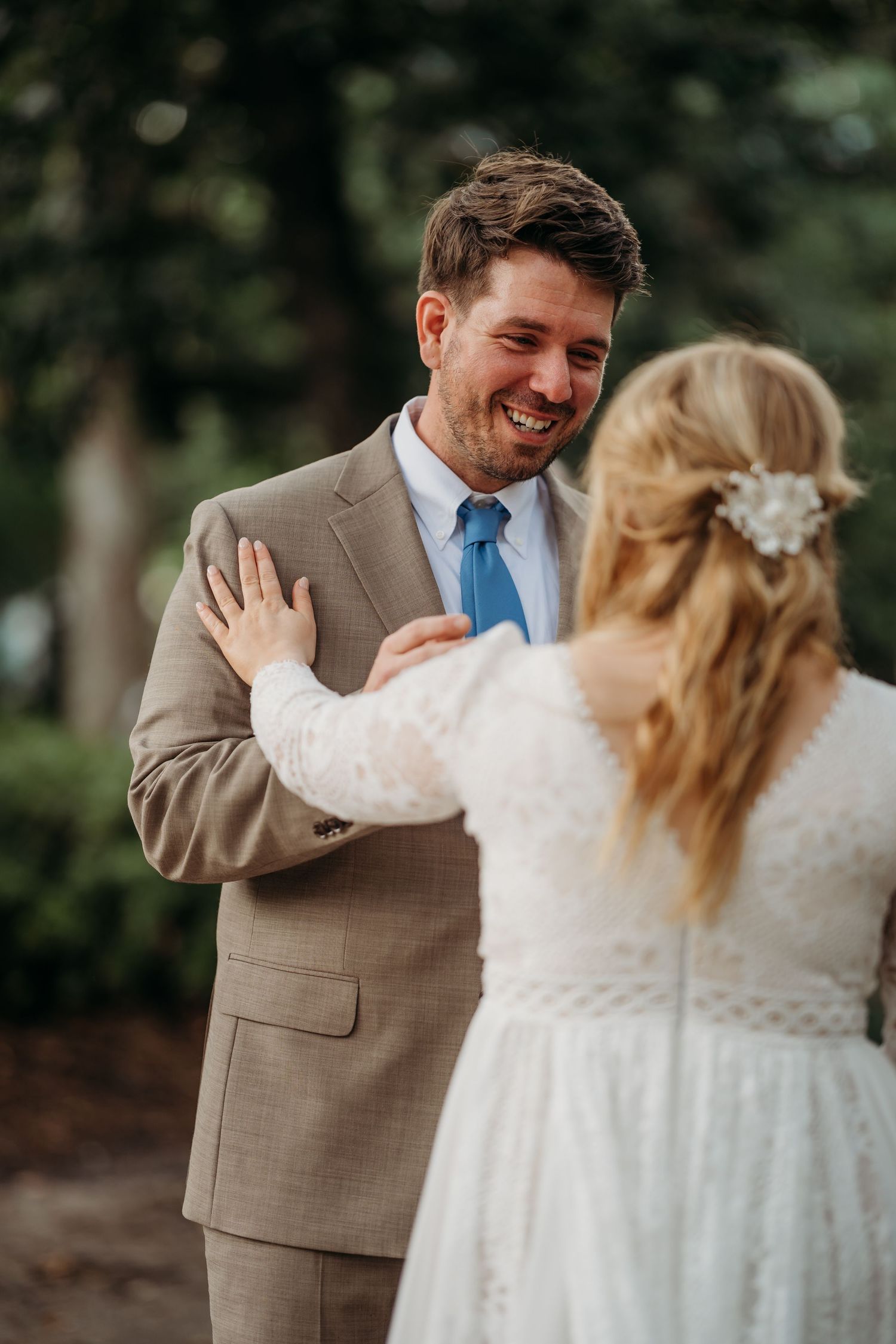 Laughing groom in tan suit and blue tie reacts joyfully as bride places hand on his chest outdoors.
