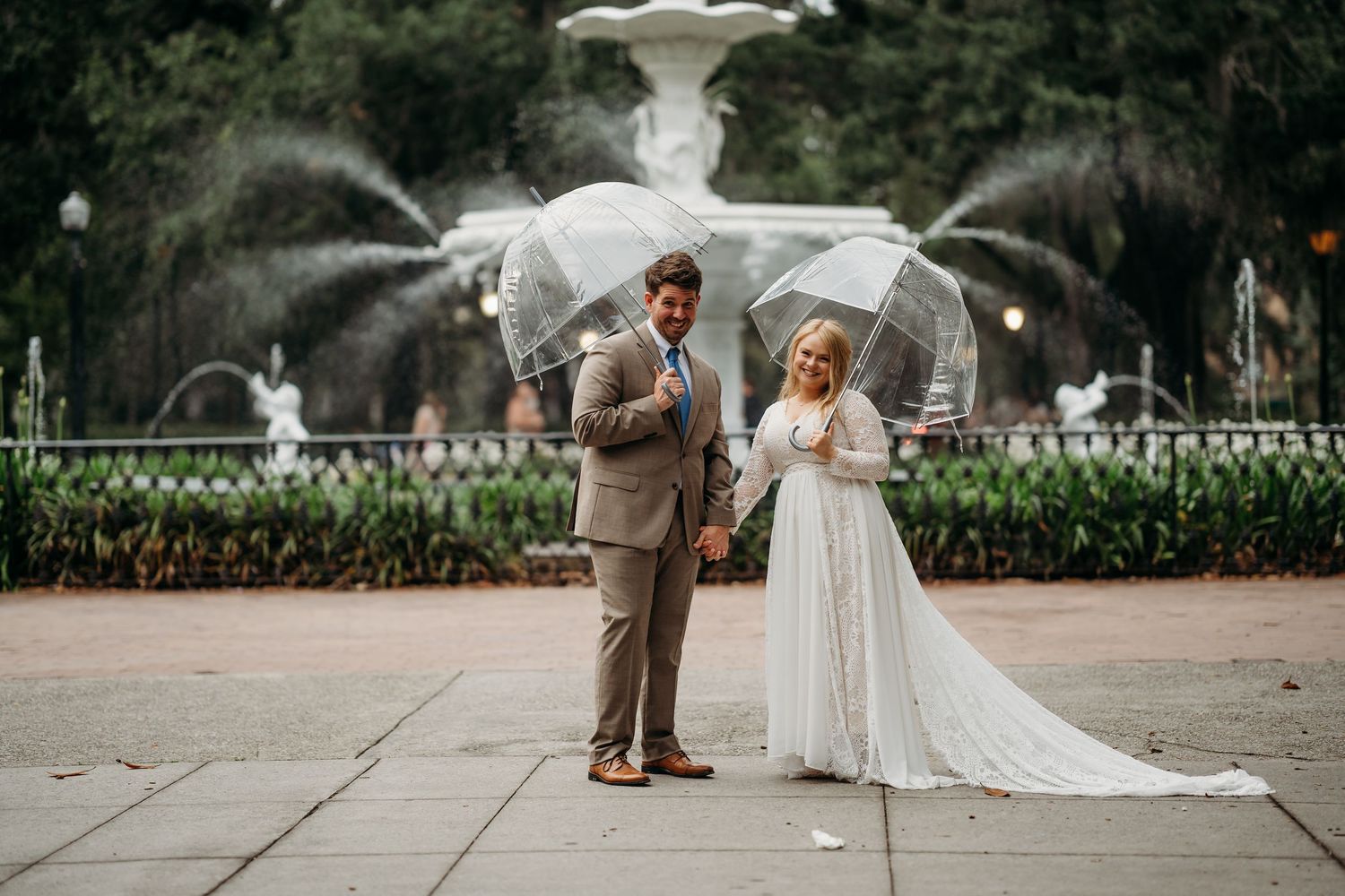 Bride and groom stand apart holding clear bubble umbrellas in front of decorative white park fountain.