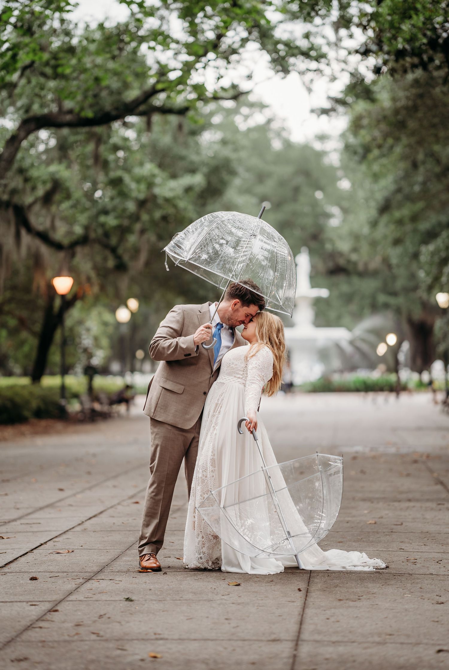 Newlyweds sharing a kiss under a clear dome umbrella near a fountain in a tree-lined park.