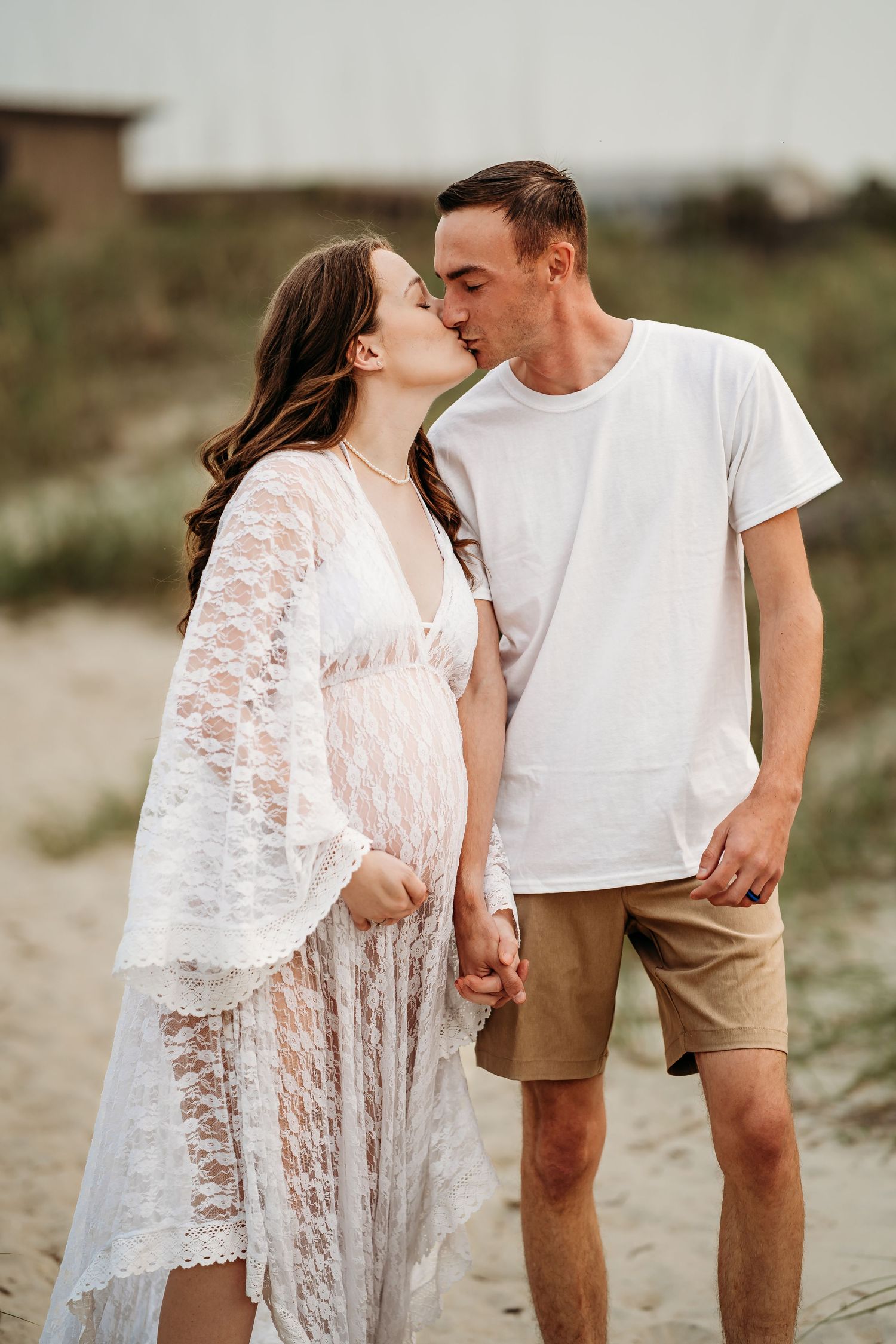Expecting couple sharing a tender kiss while holding hands on a sandy beach at dusk.