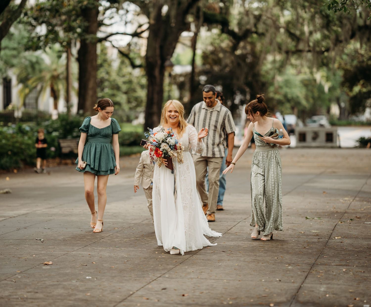 Bride in lace wedding gown walking with bridesmaids in green dresses outdoors.