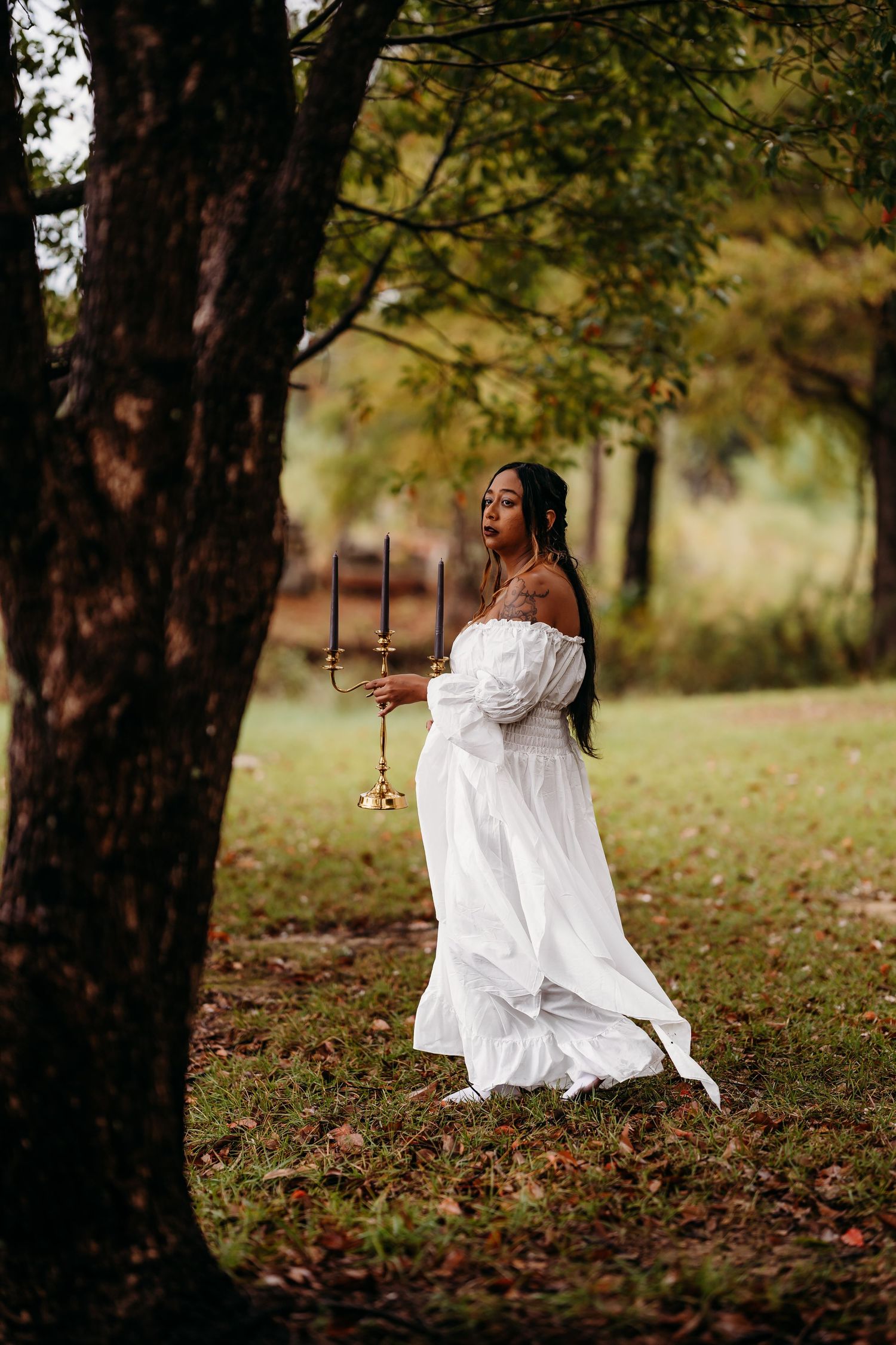 Woman in white off-shoulder dress stands under tree holding gold candelabra in autumn forest setting.