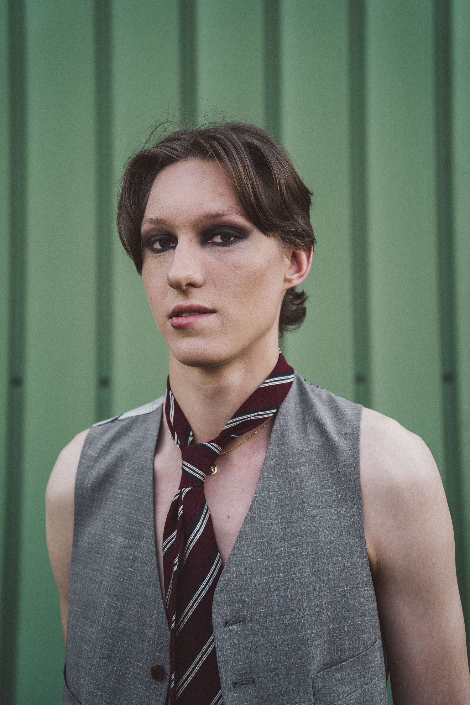 Person in gray vest and striped tie with dramatic eye makeup posing against green wall.