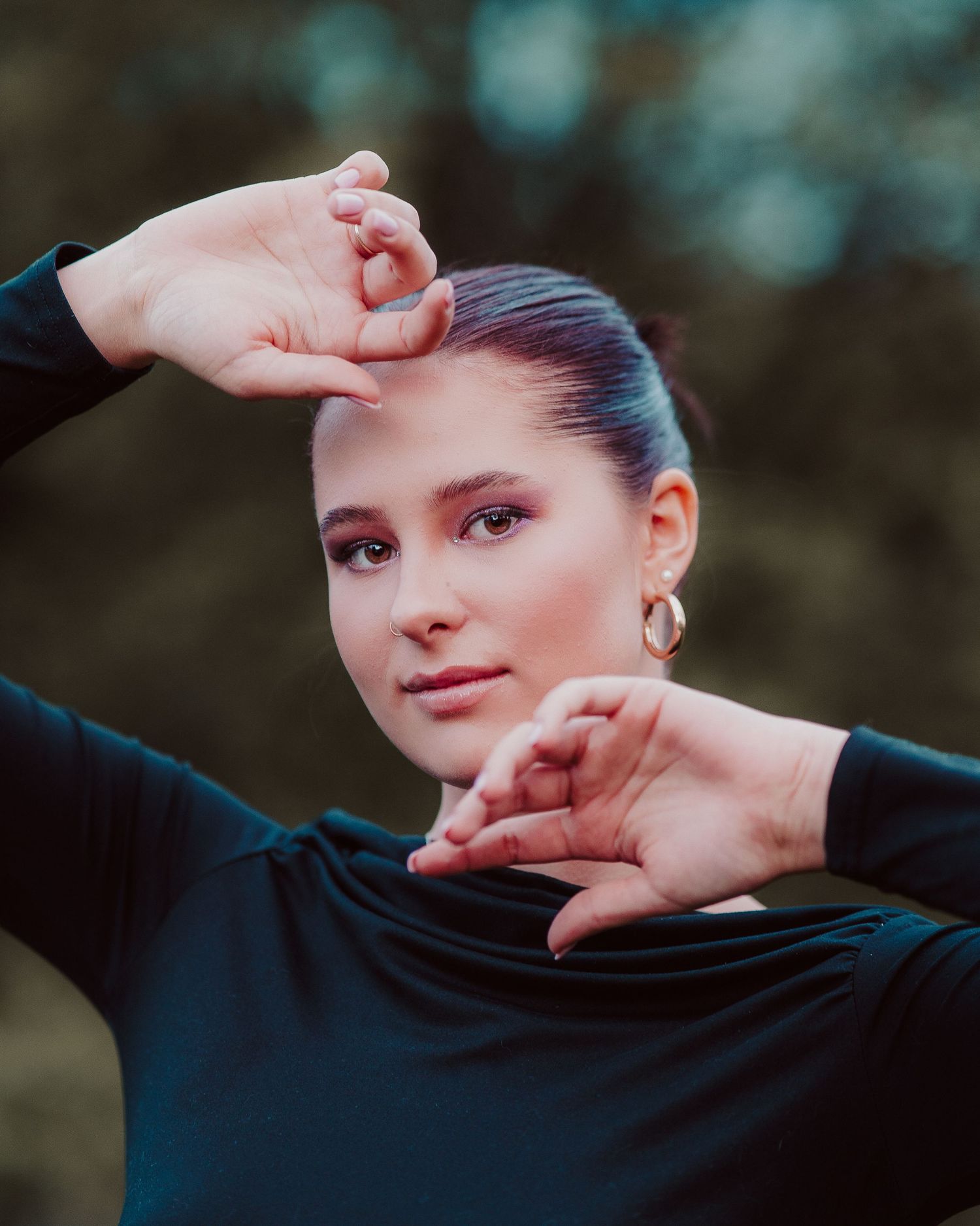 Woman in black long sleeve top posing with hands framing her face outdoors with bokeh background.