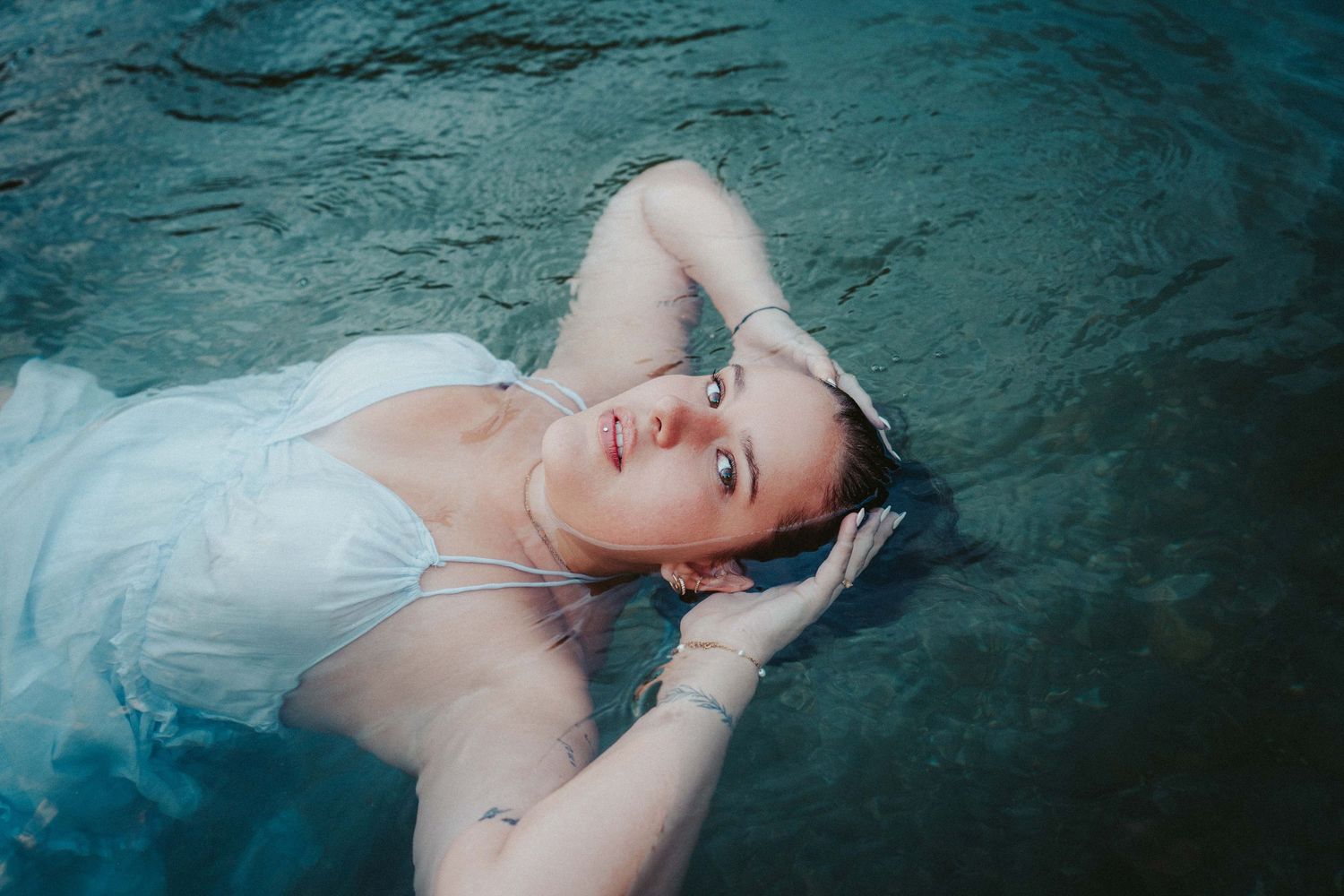 Woman in white dress floating in water with hands behind head, ethereal photography.