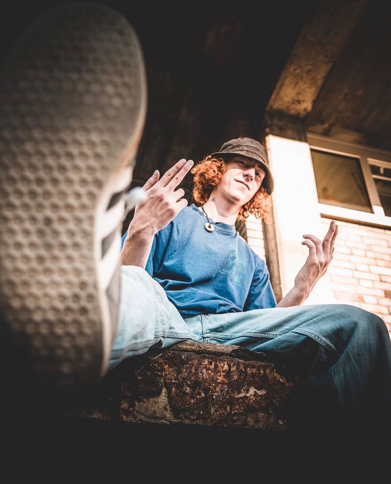 Curly redhead in blue shirt and bucket hat sitting on ledge photographed from low angle perspective.
