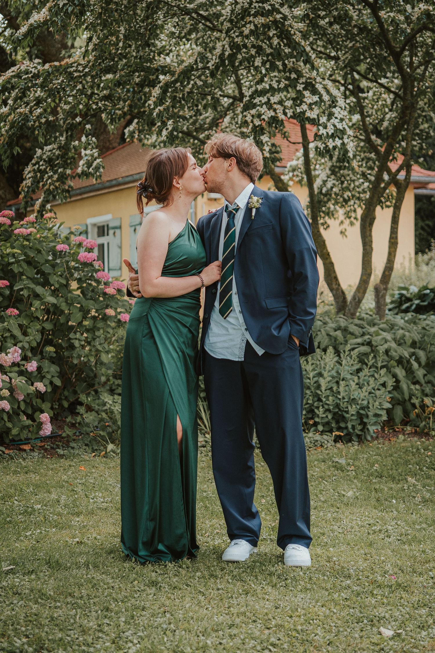 Couple sharing a romantic kiss outdoors, wearing elegant green gown and navy suit with boutonniere in a garden setting.