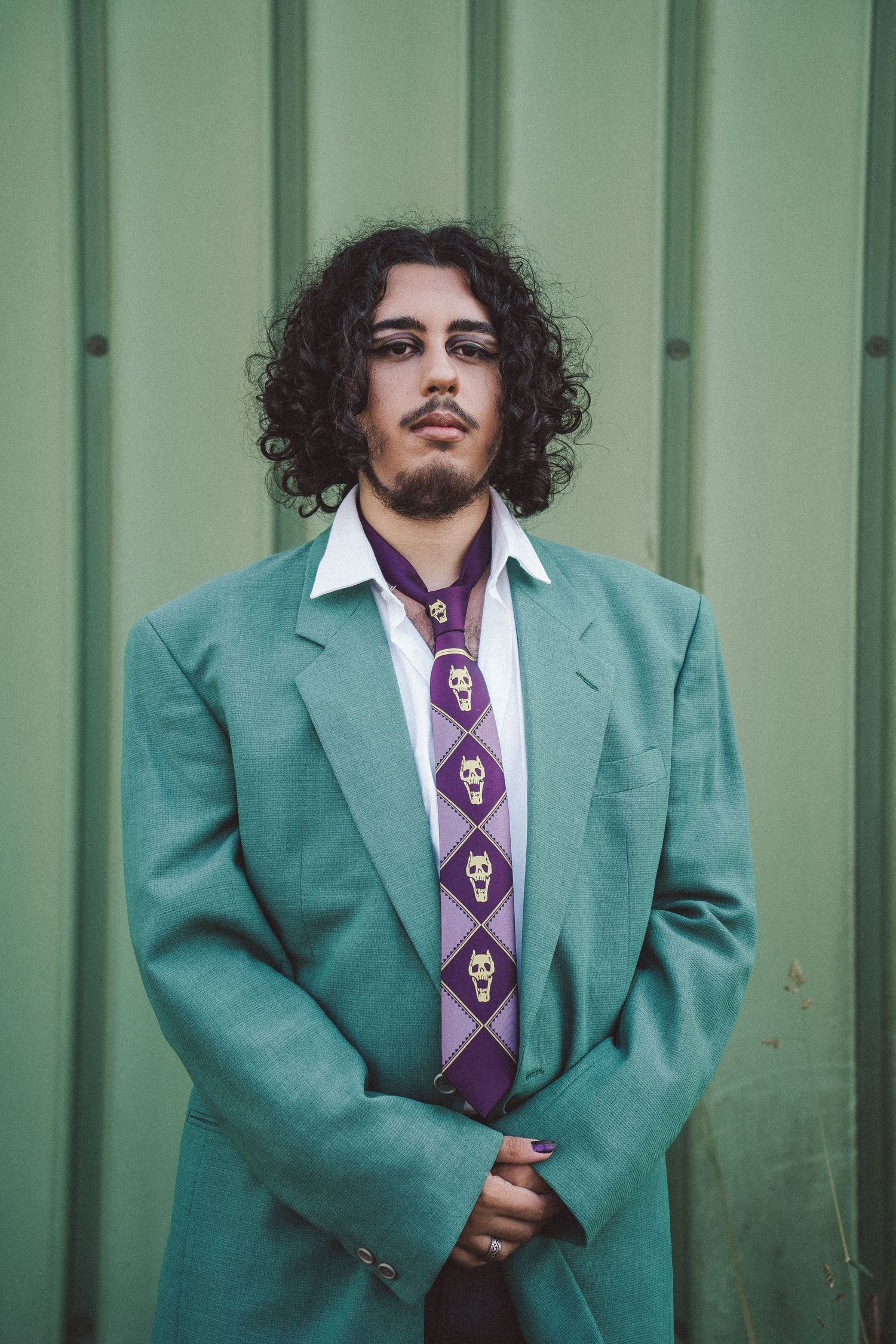 Person with dark curly hair in teal suit and skull tie posing against green wall.