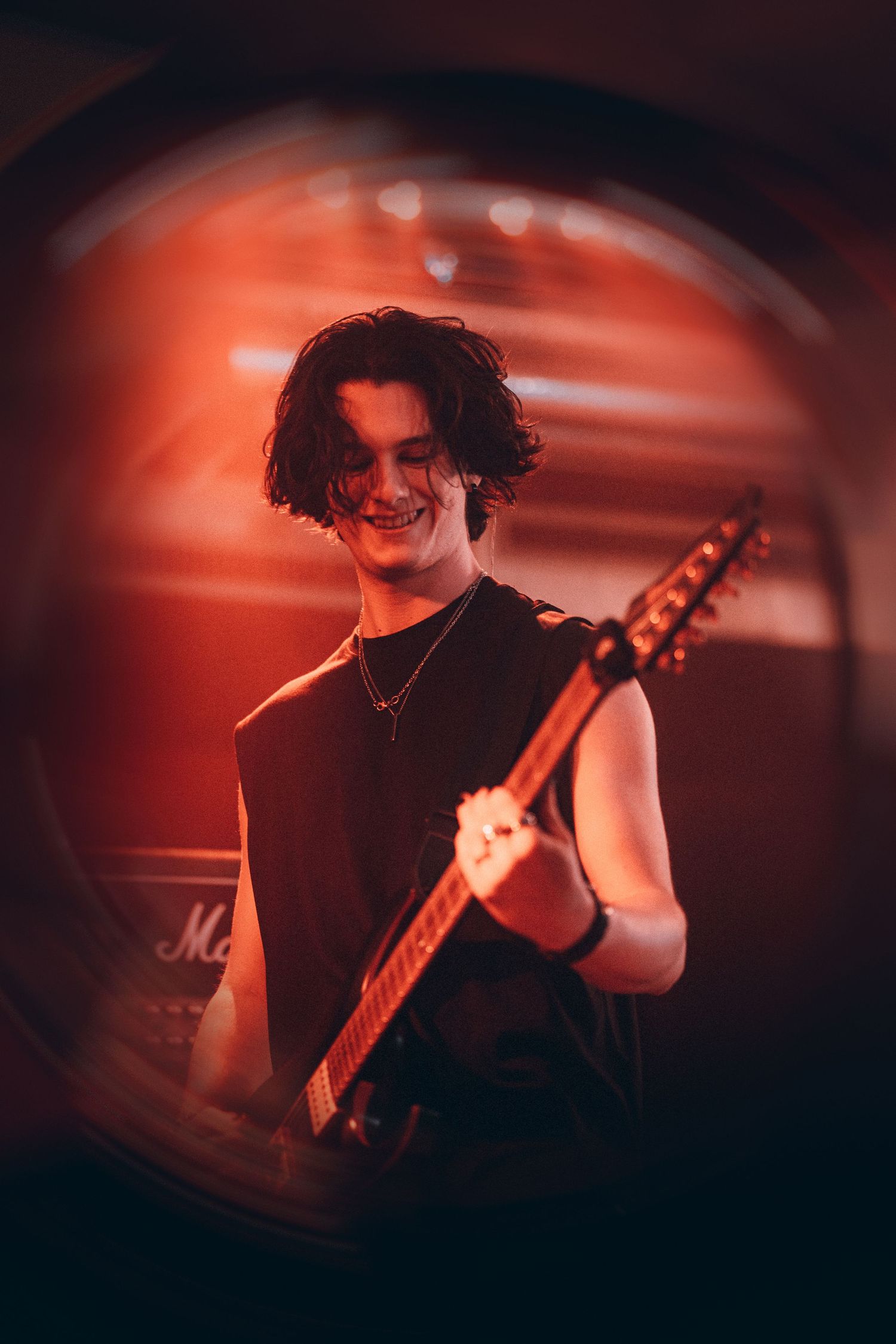 Smiling guitarist with dark hair playing electric guitar under red stage lights.