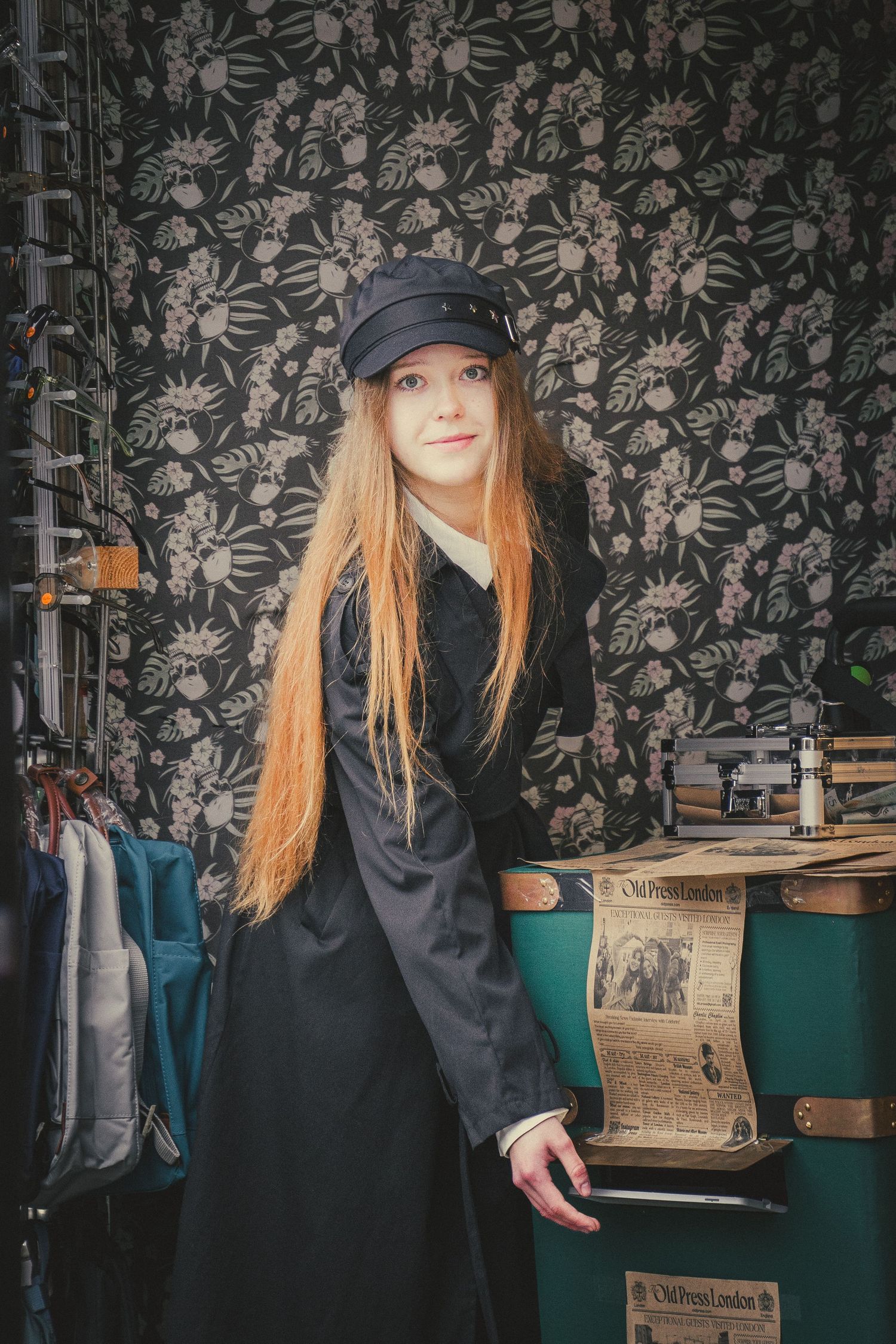 Young woman with long hair in a black coat and cap poses in a vintage-style market stall.